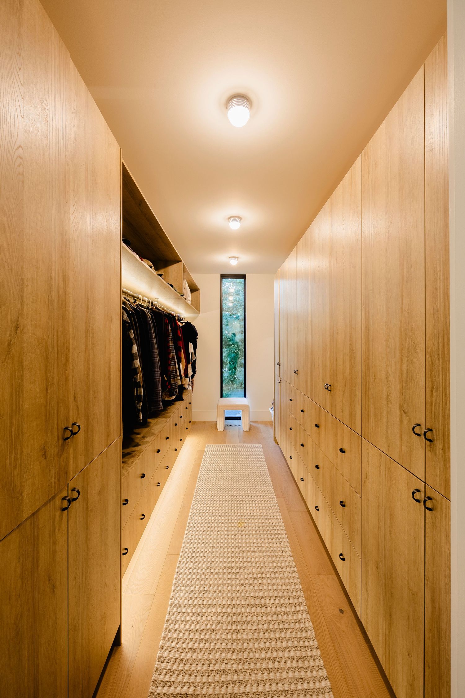 Spacious walk-in closet with floor-to-ceiling wood cabinetry, runner rug, and narrow vertical window.