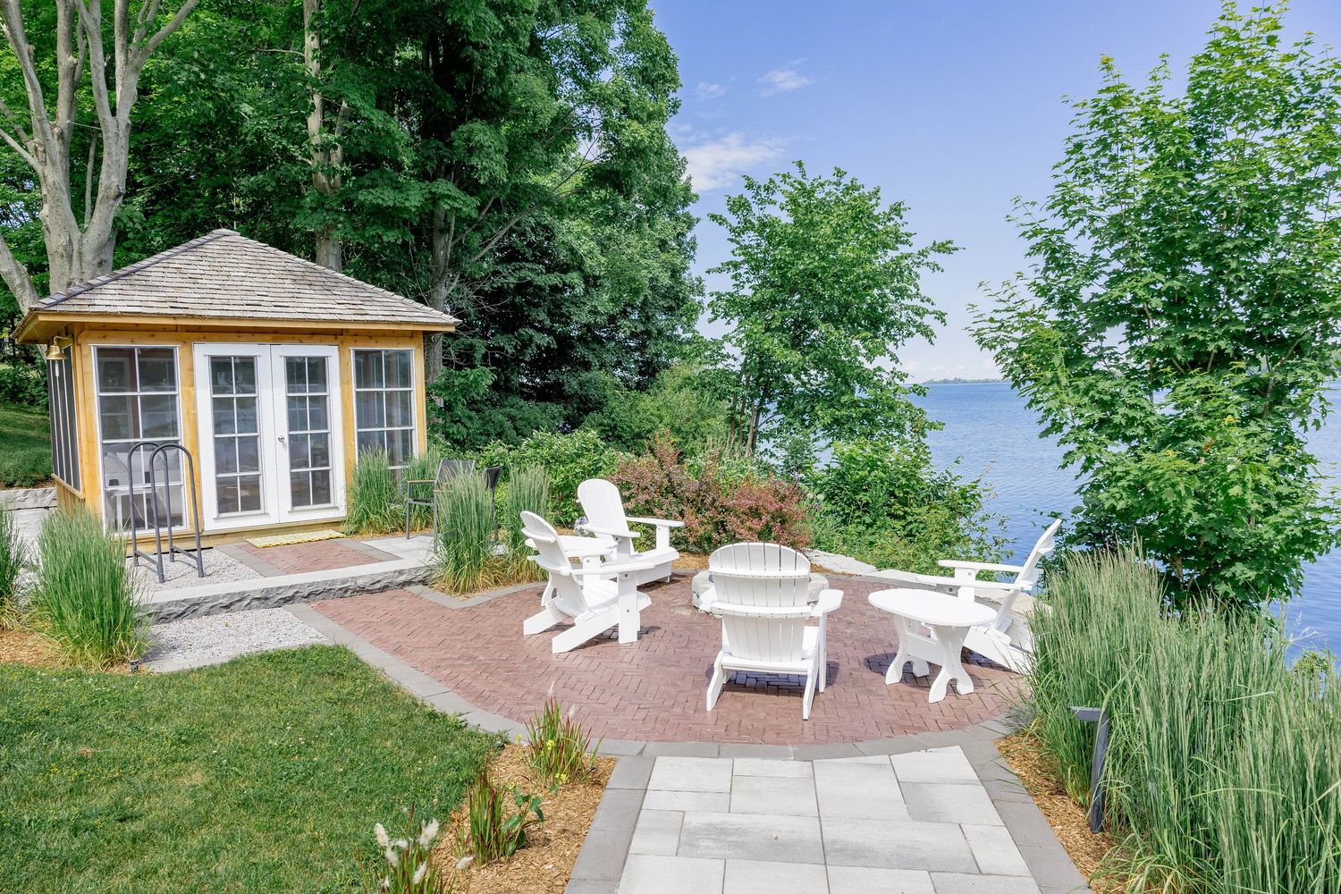 Charming yellow garden shed with white Adirondack chairs on a brick patio overlooking a lake.