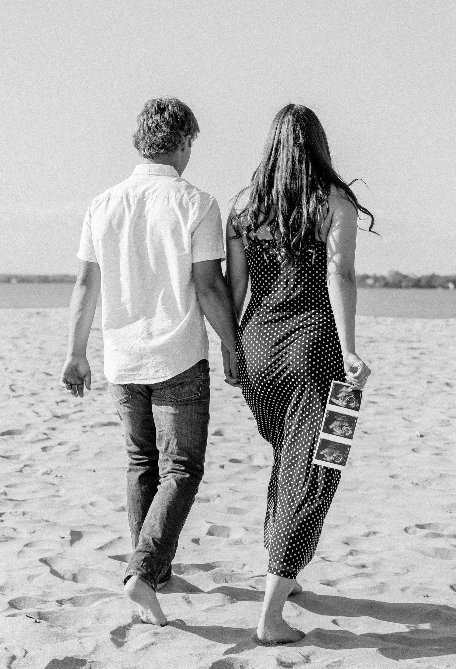 Black and white photo of a couple walking on the beach holding hands, woman carrying ultrasound photos.