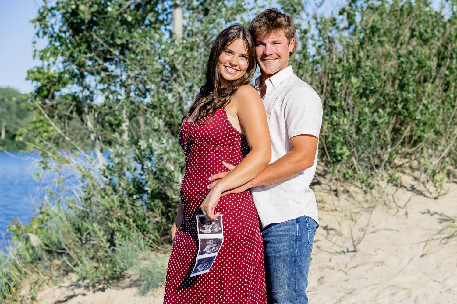 Smiling pregnant couple holding ultrasound photos on sandy beach with trees and river in background on sunny day.