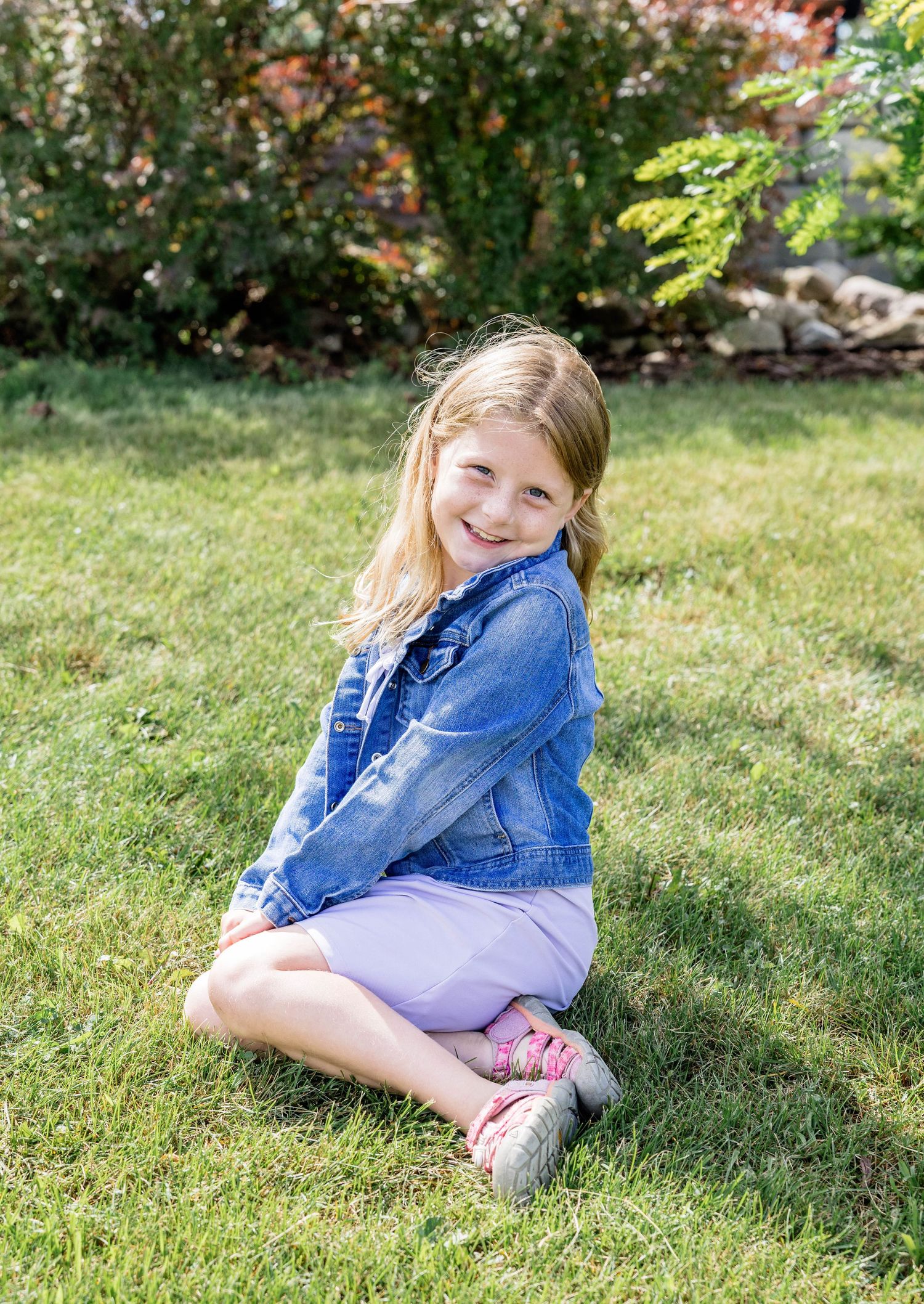 Young girl in denim jacket and purple skirt sitting cross-legged on green grass, smiling outdoors.