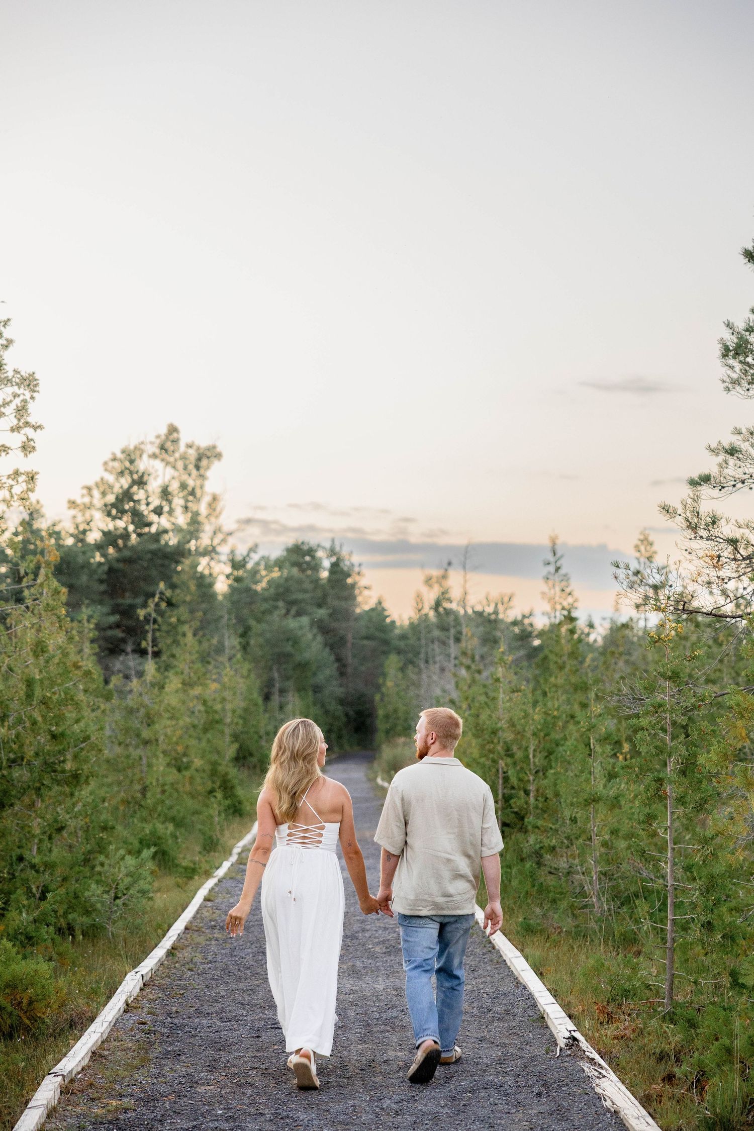 Couple holding hands walking down a tree-lined path at sunset, woman in white dress and man in casual attire.