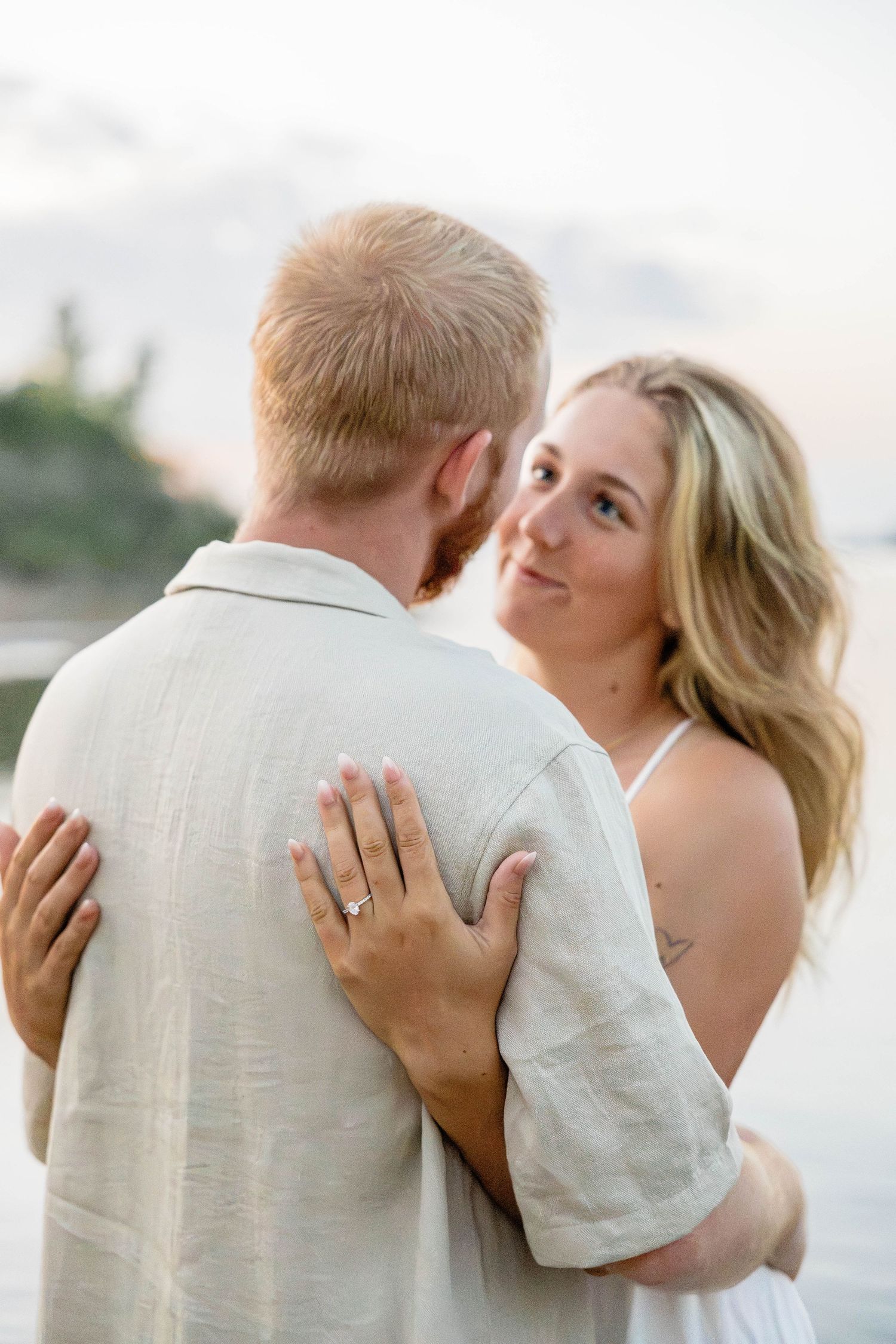 Couple embracing on beach at sunset, woman in white dress with engagement ring smiling over mans shoulder.
