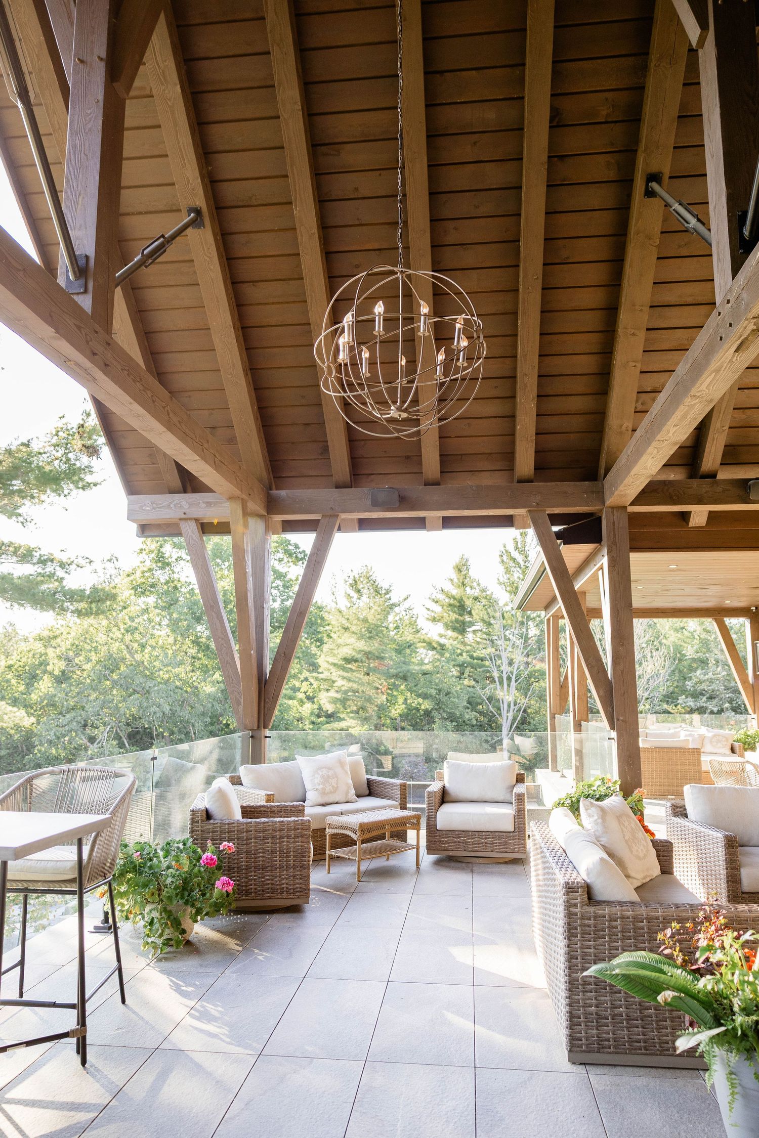 Covered outdoor patio with timber frame roof, globe chandelier, wicker furniture, and garden views.
