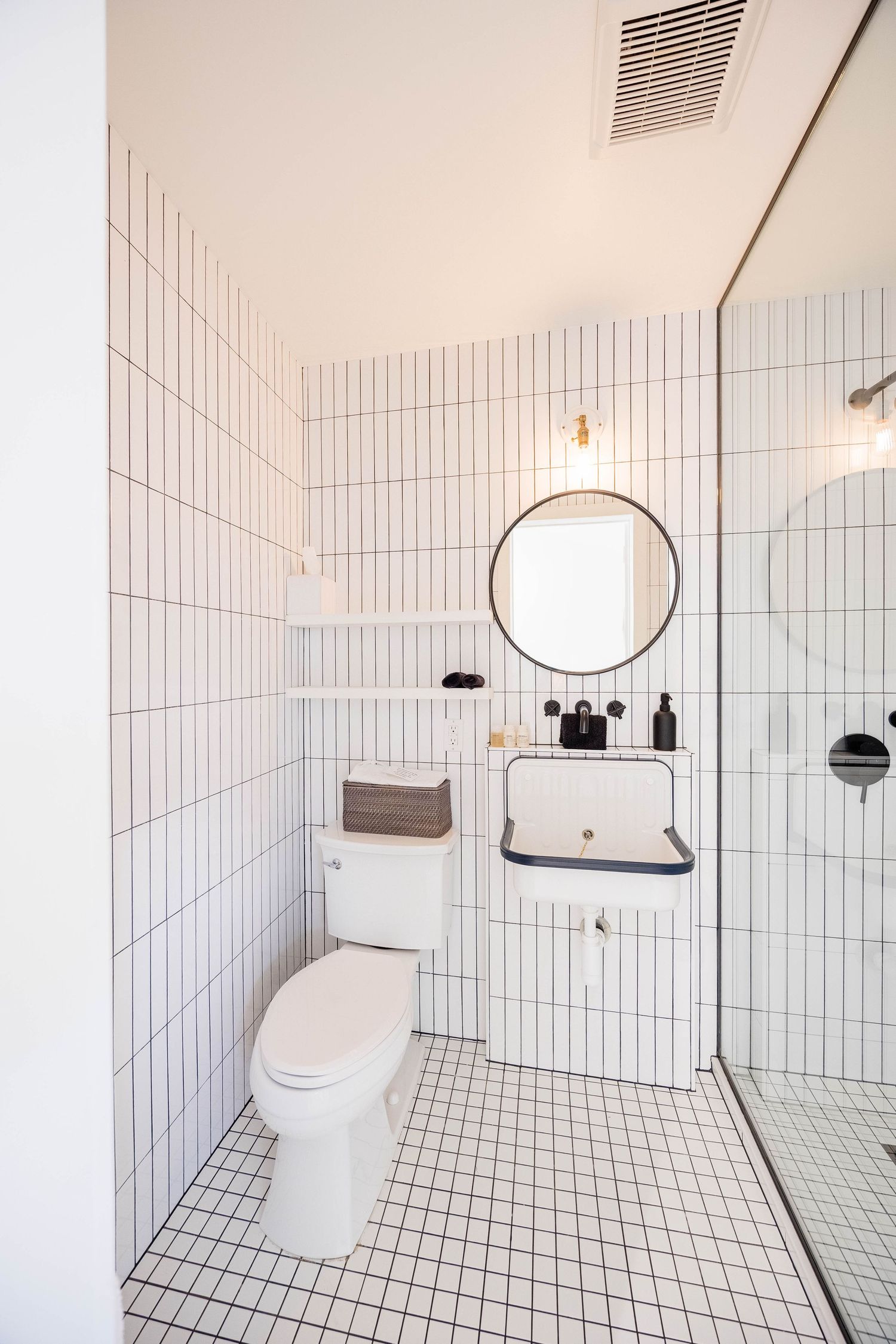 Clean white tiled bathroom with toilet, wall-mounted sink, round mirror, and glass shower.