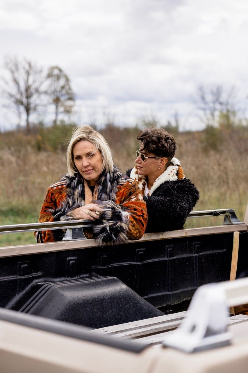 Two fashionable women leaning on the side of a vintage pickup truck bed in an open field.