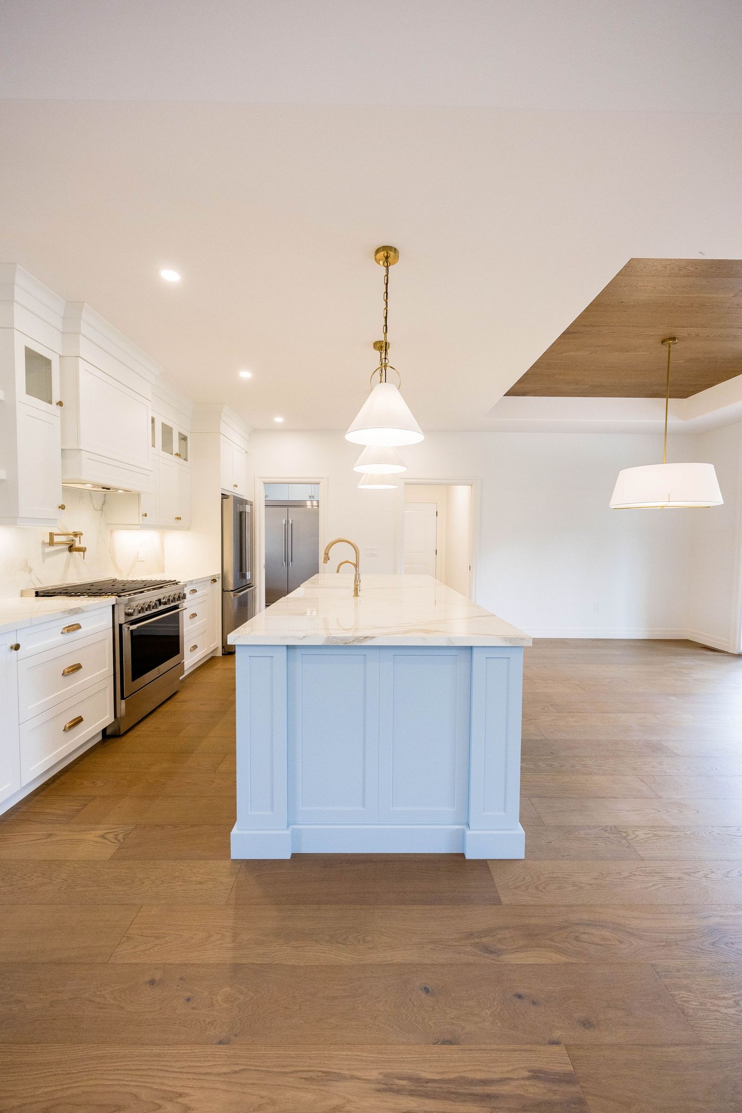 Modern white kitchen with large marble island, hardwood floors, pendant lights, and stainless steel appliances.