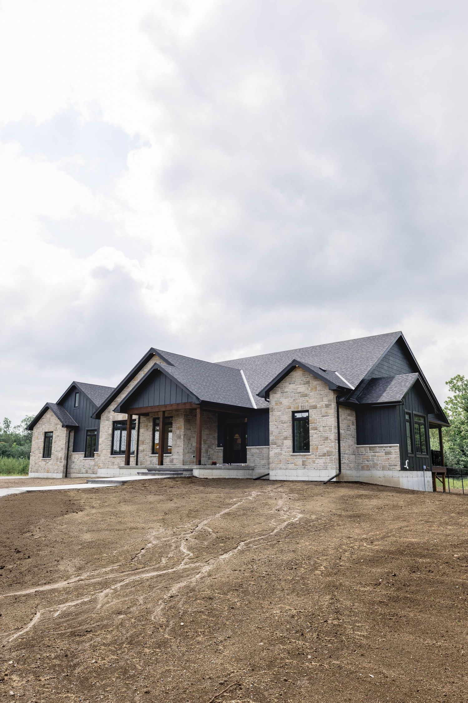 Modern farmhouse with dark siding, stone exterior, and gray roof under cloudy skies on a freshly graded dirt lot.