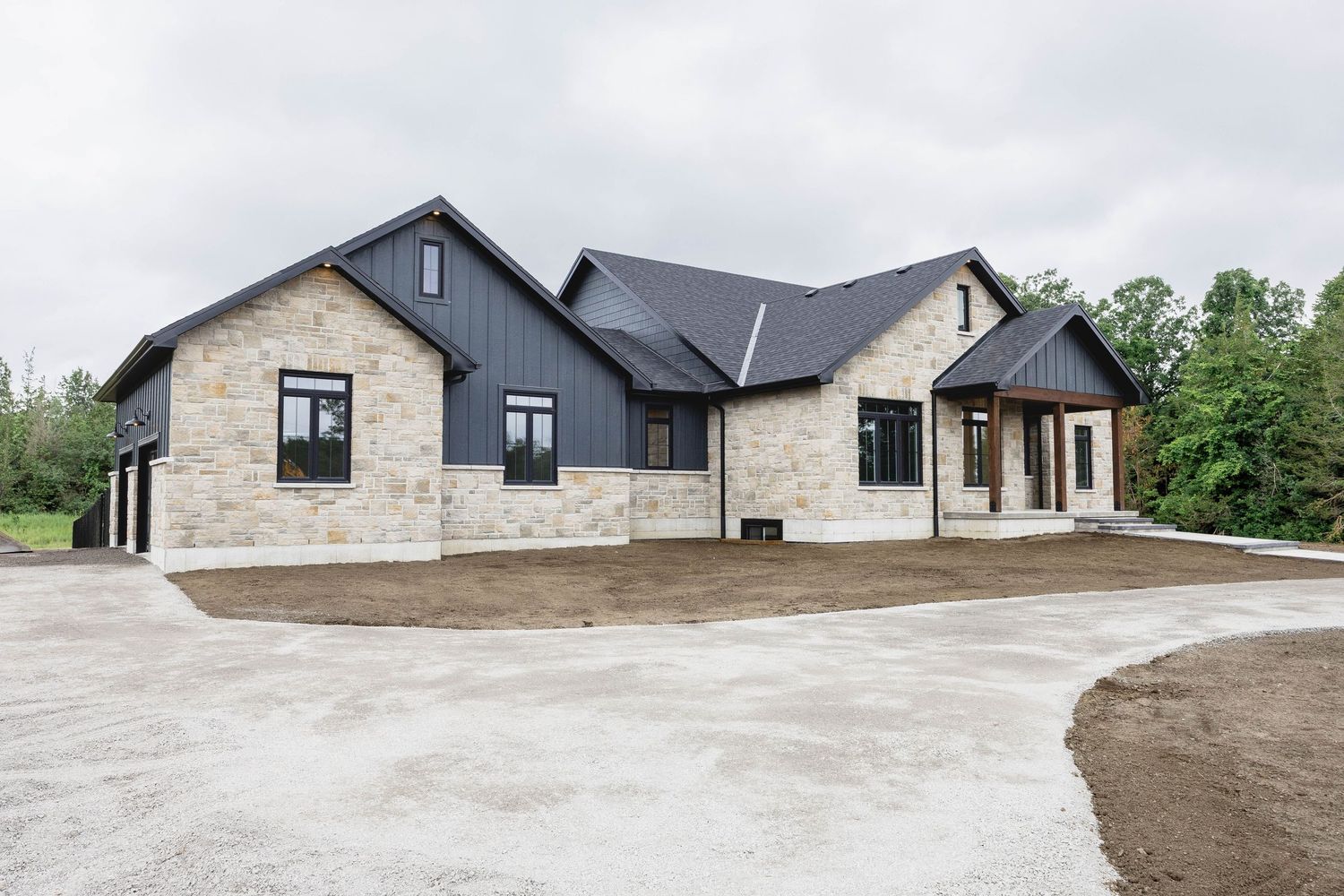 Modern farmhouse with stone and dark siding, black roof, and circular driveway surrounded by trees.