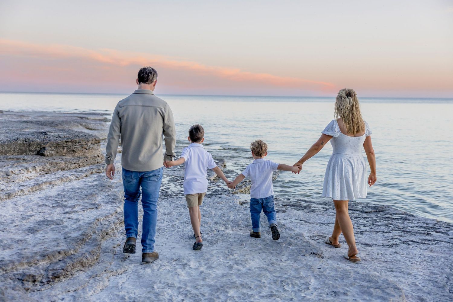 Family of four holding hands walking along a scenic beach at sunset.