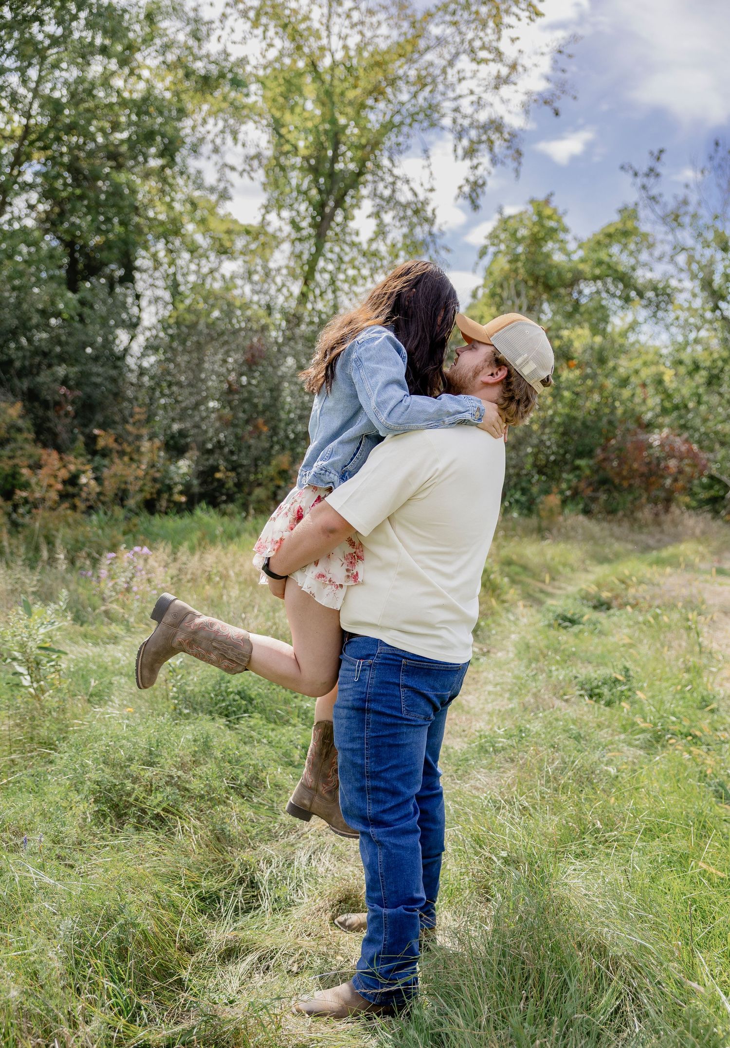 A man lifts and kisses a woman in a sunny green meadow, both wearing casual country-style outfits.