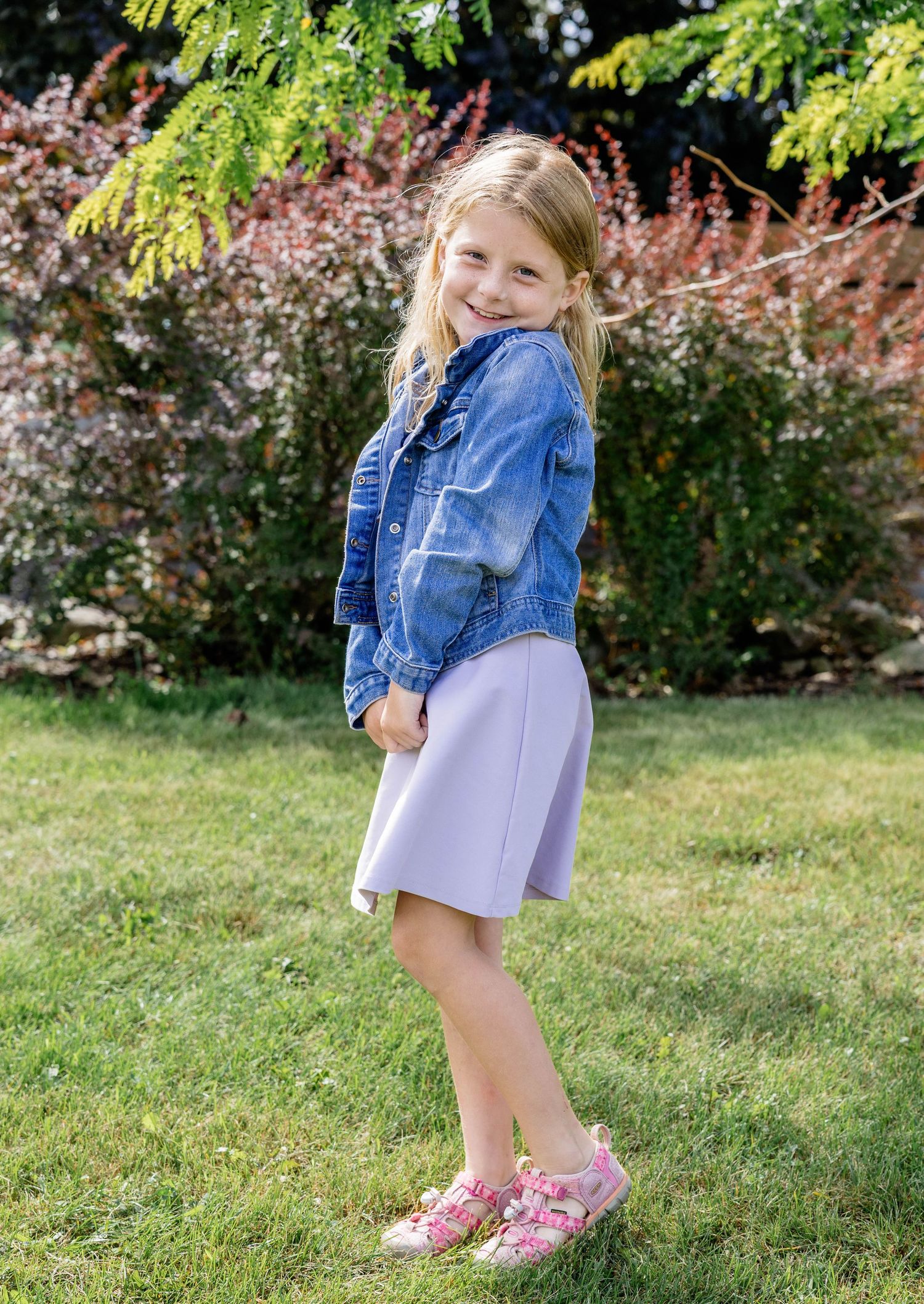 Young girl in denim jacket and lavender skirt smiling outdoors on green grass with lush garden background.