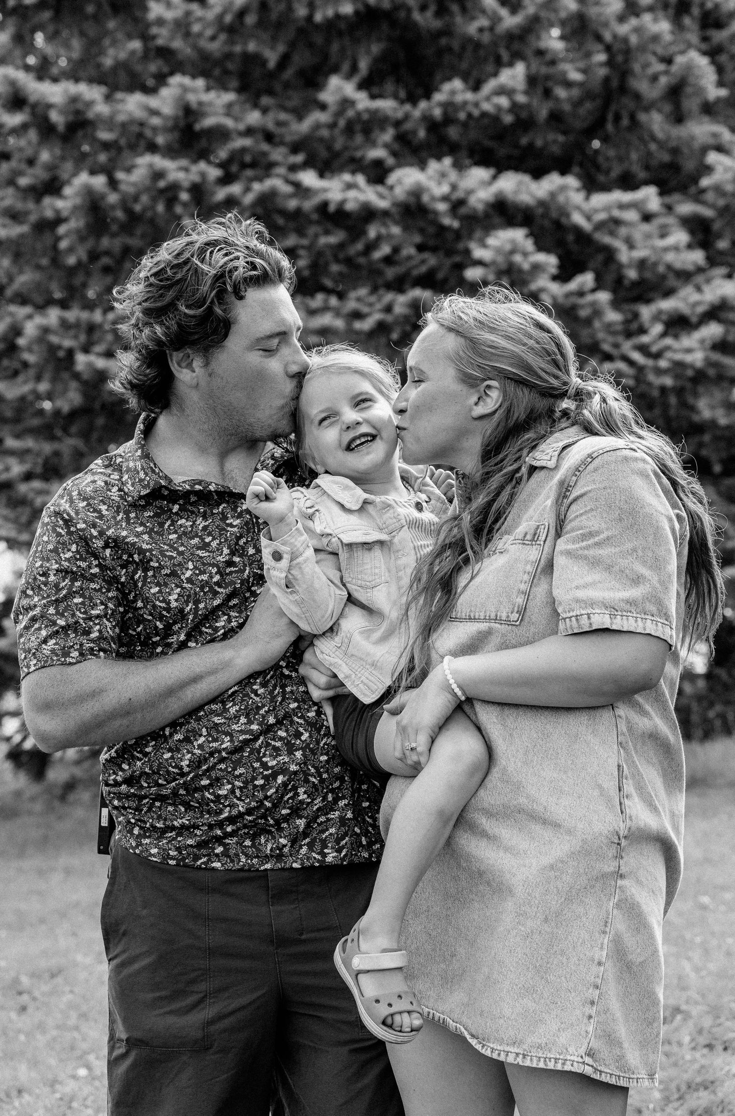 Black and white photo of parents kissing their laughing toddler on the cheeks outdoors near trees.