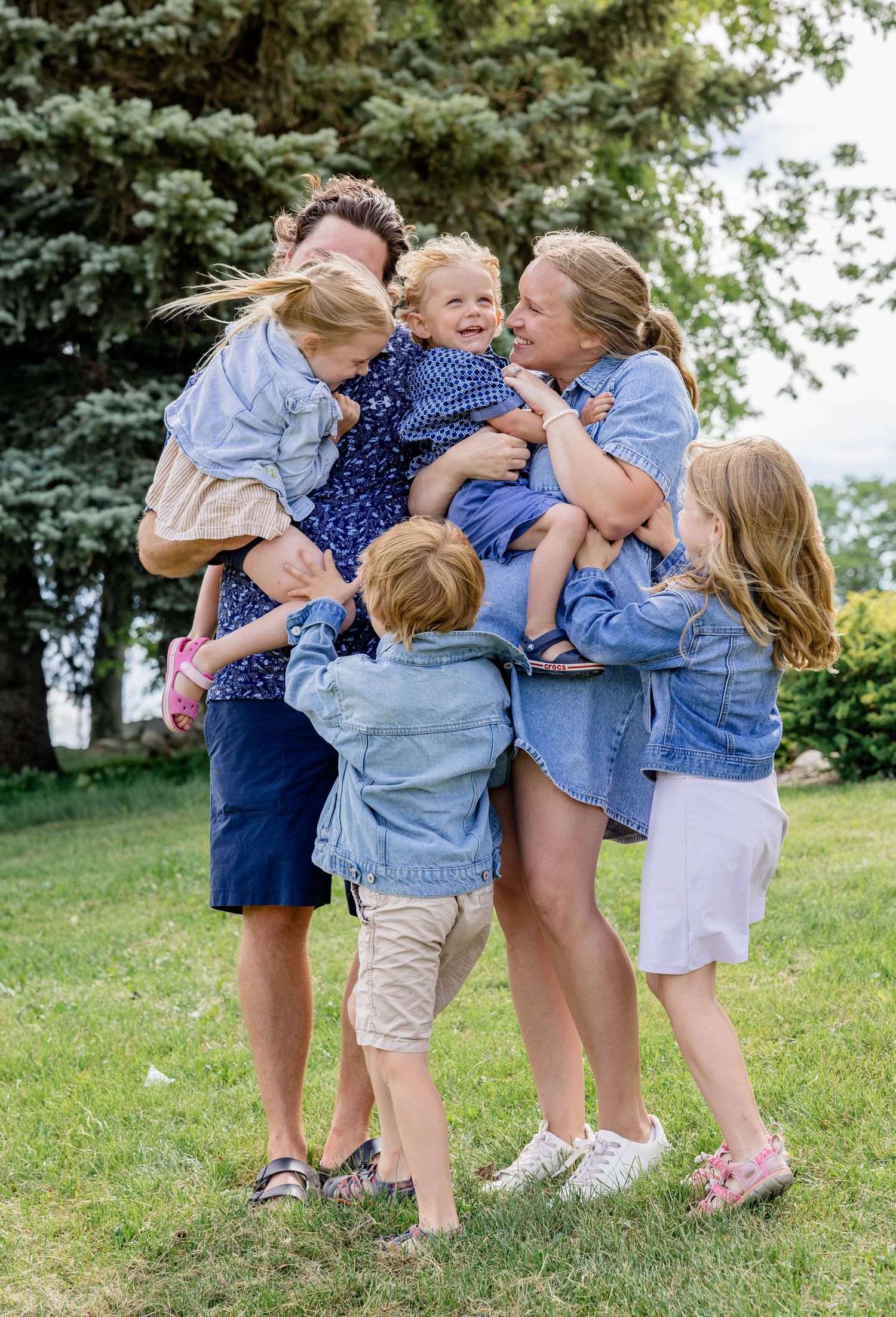 A joyful family in coordinating blue outfits hugging outdoors on green grass with trees in the background.