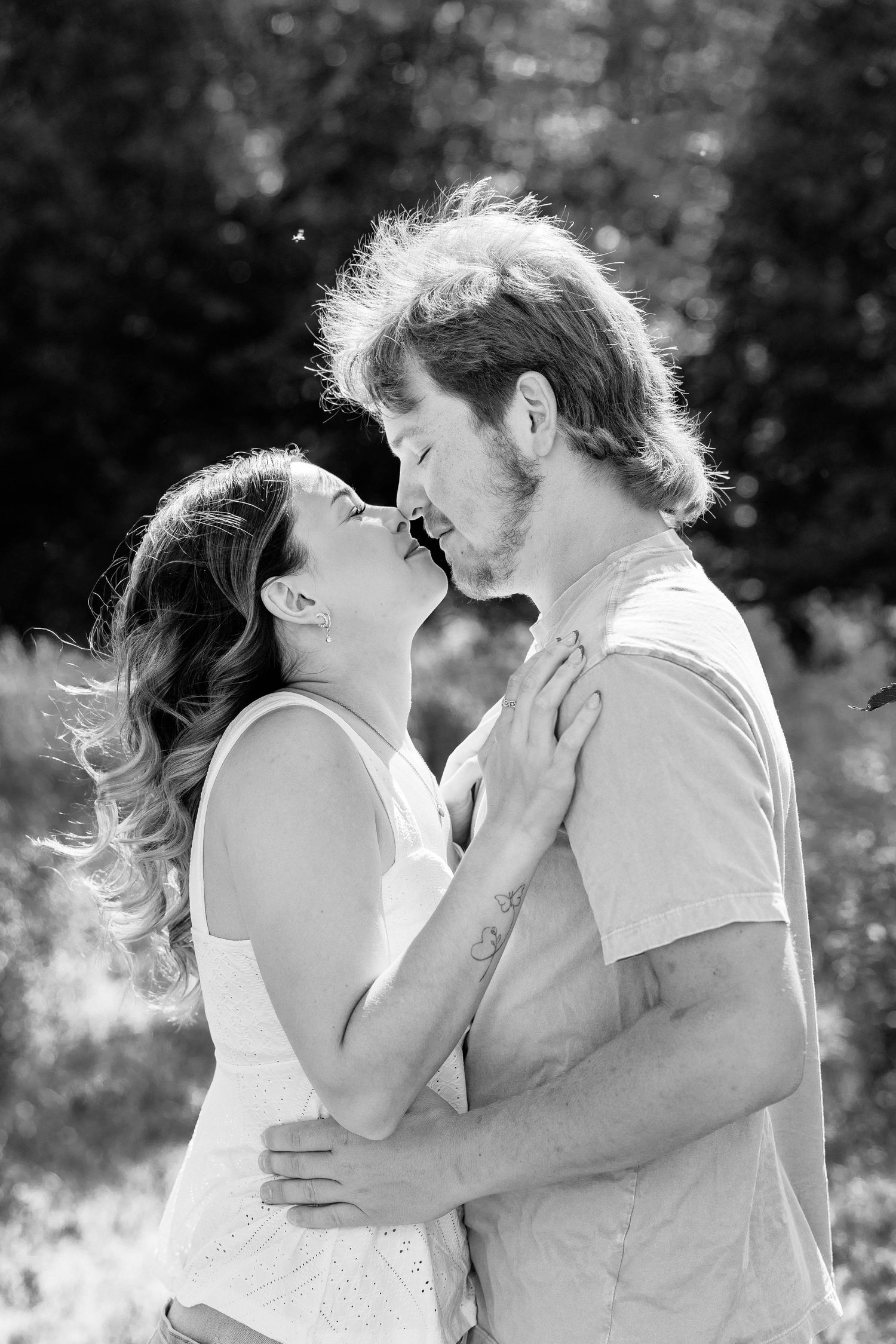 Black and white photo of a couple sharing a romantic kiss outdoors, embracing in a sun-dappled natural setting.