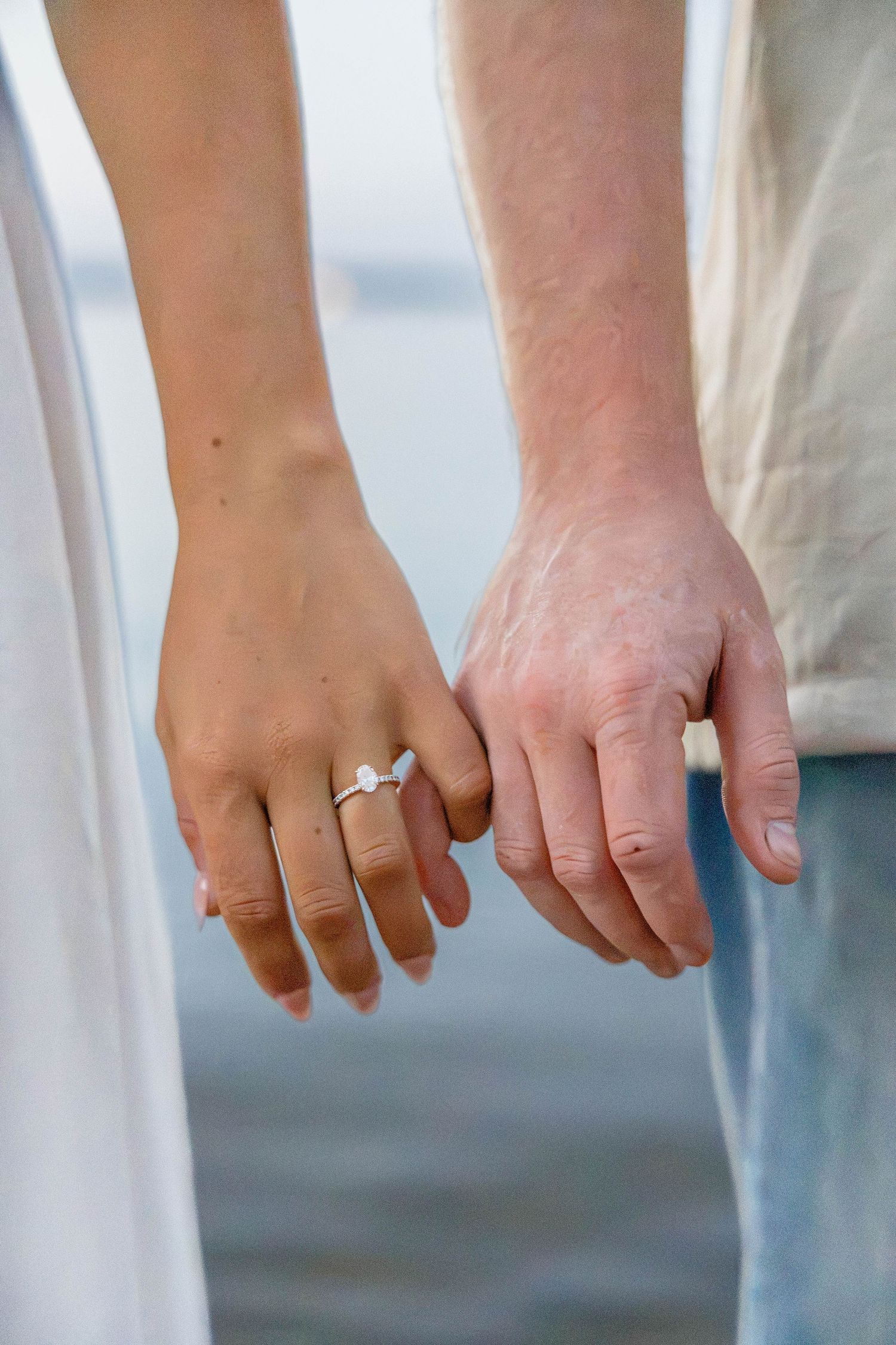 Close-up of a couple holding hands outdoors near water, woman wearing a diamond engagement ring on her finger.