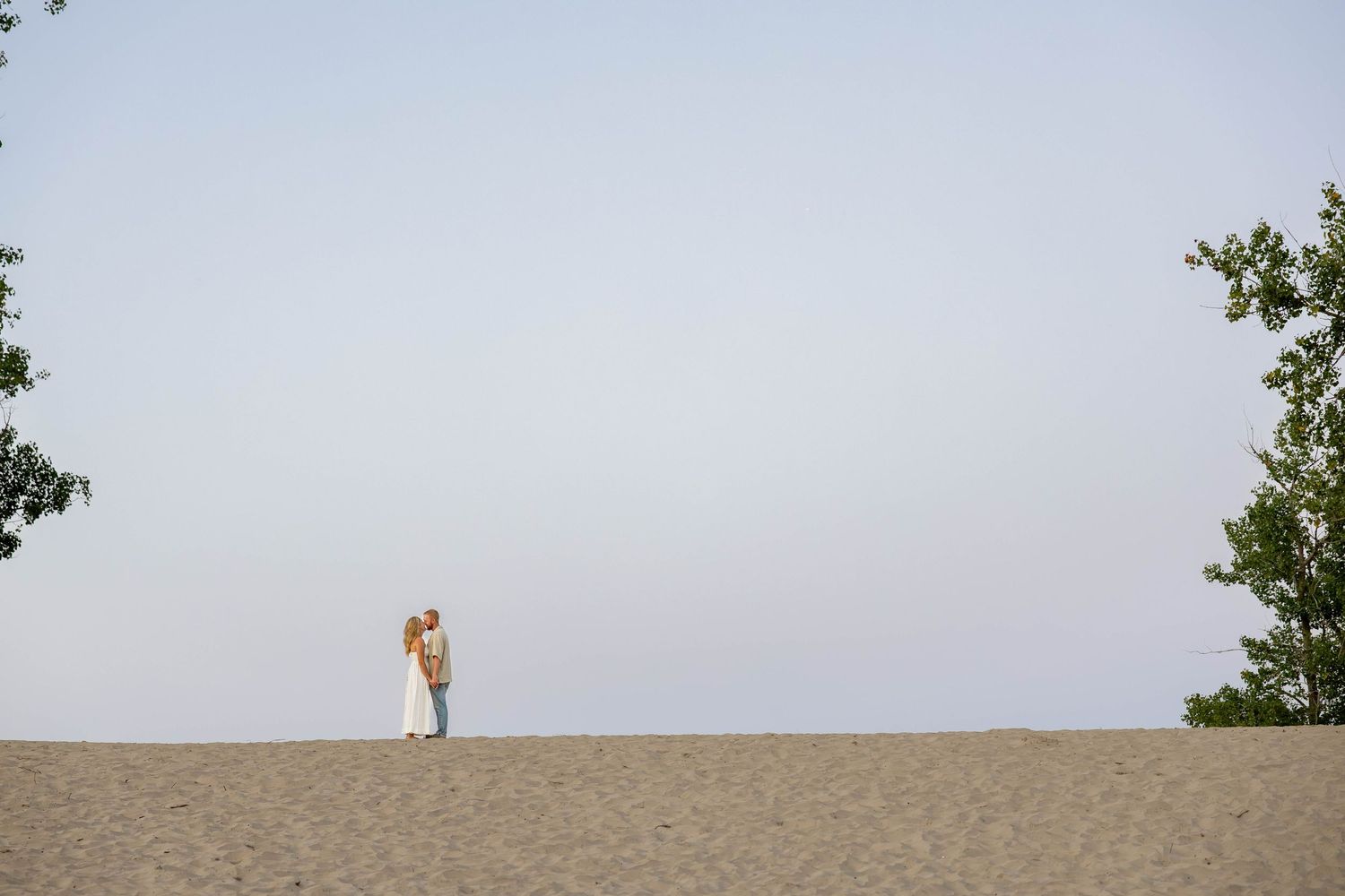 A couple shares a romantic kiss on a sandy beach dune, framed by green trees against a clear sky.