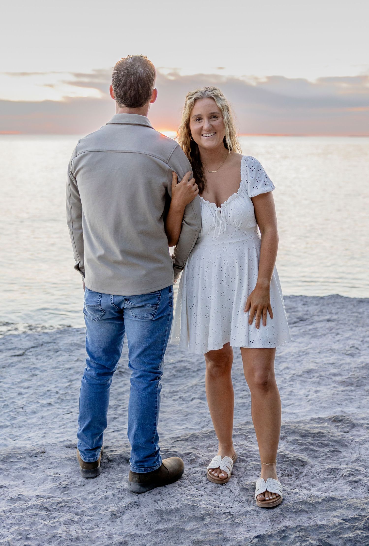 Couple standing on rocky beach at sunset, woman in white dress smiling at camera.