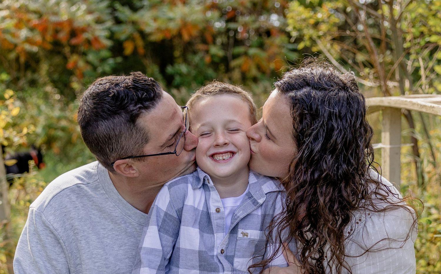 Parents kissing their smiling young boy on both cheeks outdoors with green foliage in the background.