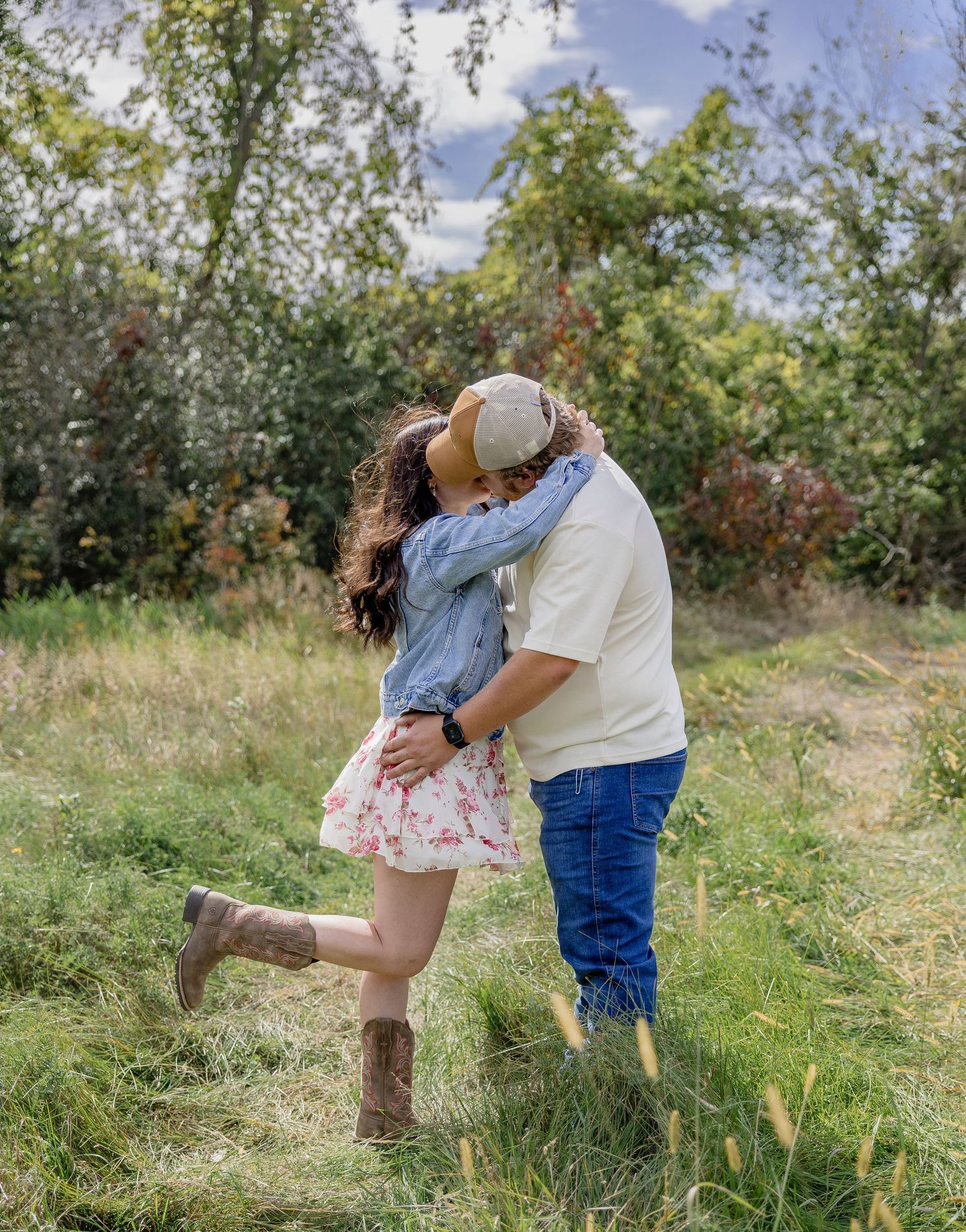 Couple kissing in a grassy field, woman in floral skirt and cowboy boots, man in jeans and tan hat, trees in background.