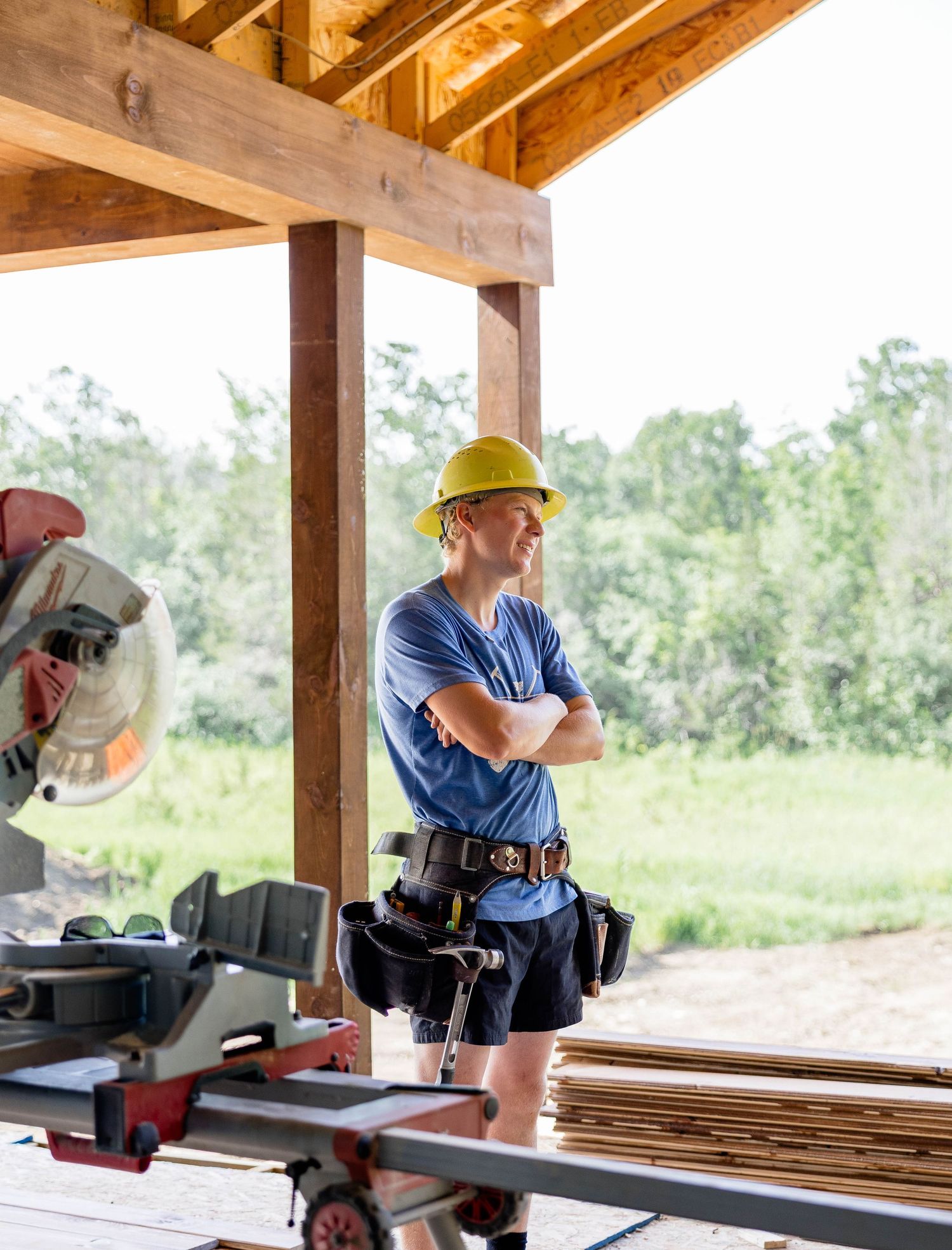 Construction worker in yellow hard hat standing with arms crossed at a wood-framed building site with power saw.
