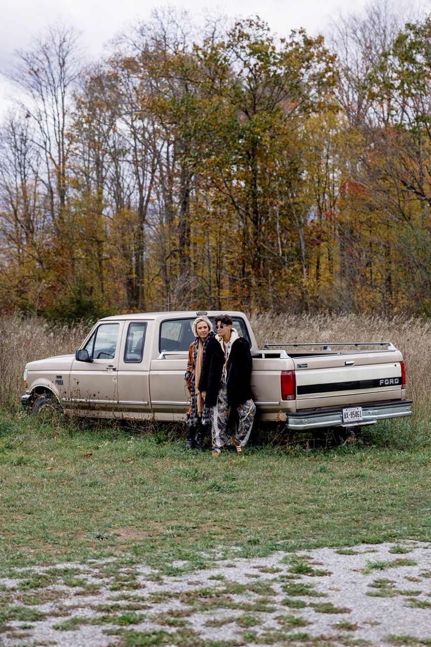 Two women in bohemian coats standing beside a vintage beige Ford pickup truck in a fall field.
