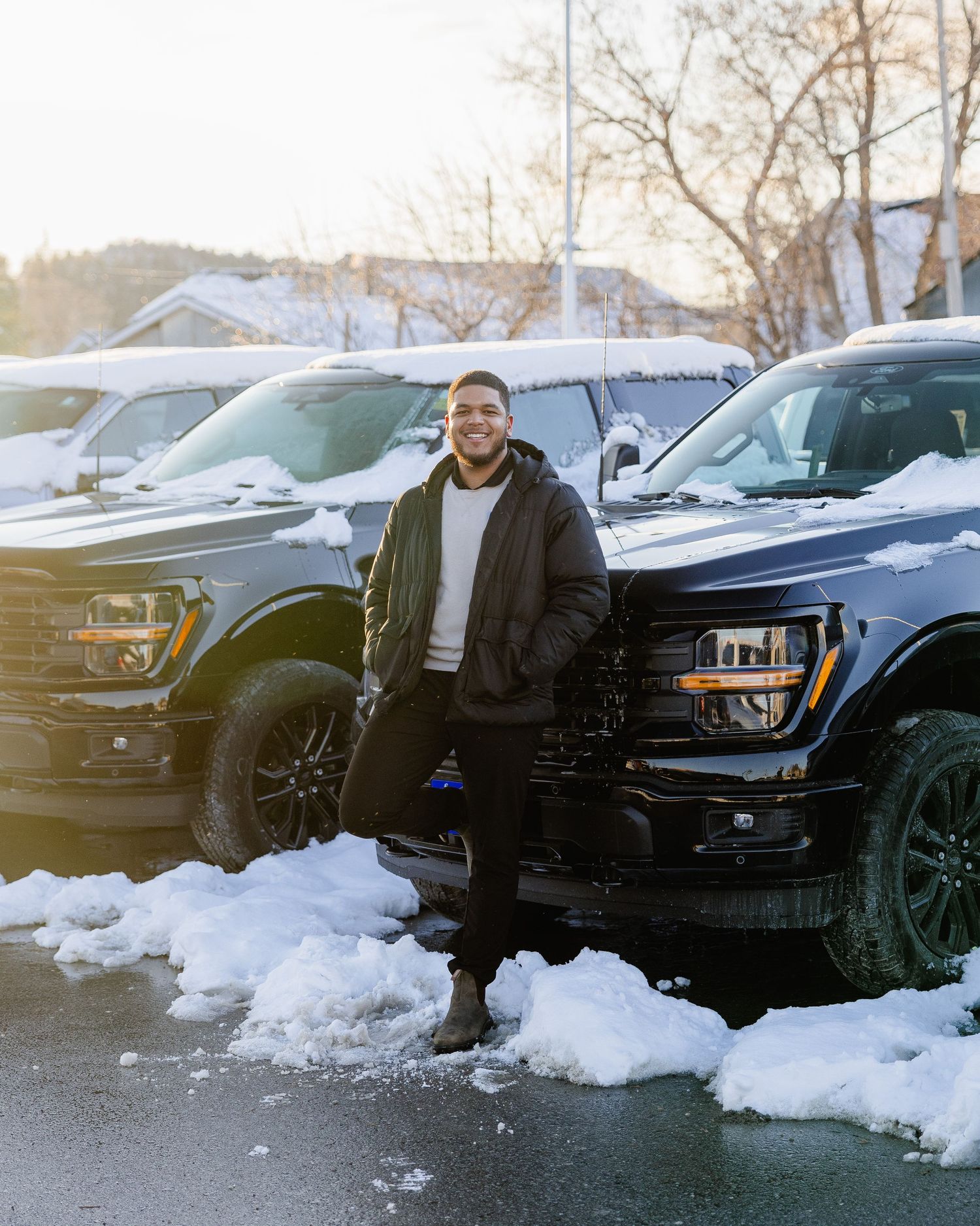 Man standing beside dark Ford Raptor trucks in snowy parking lot at golden hour.