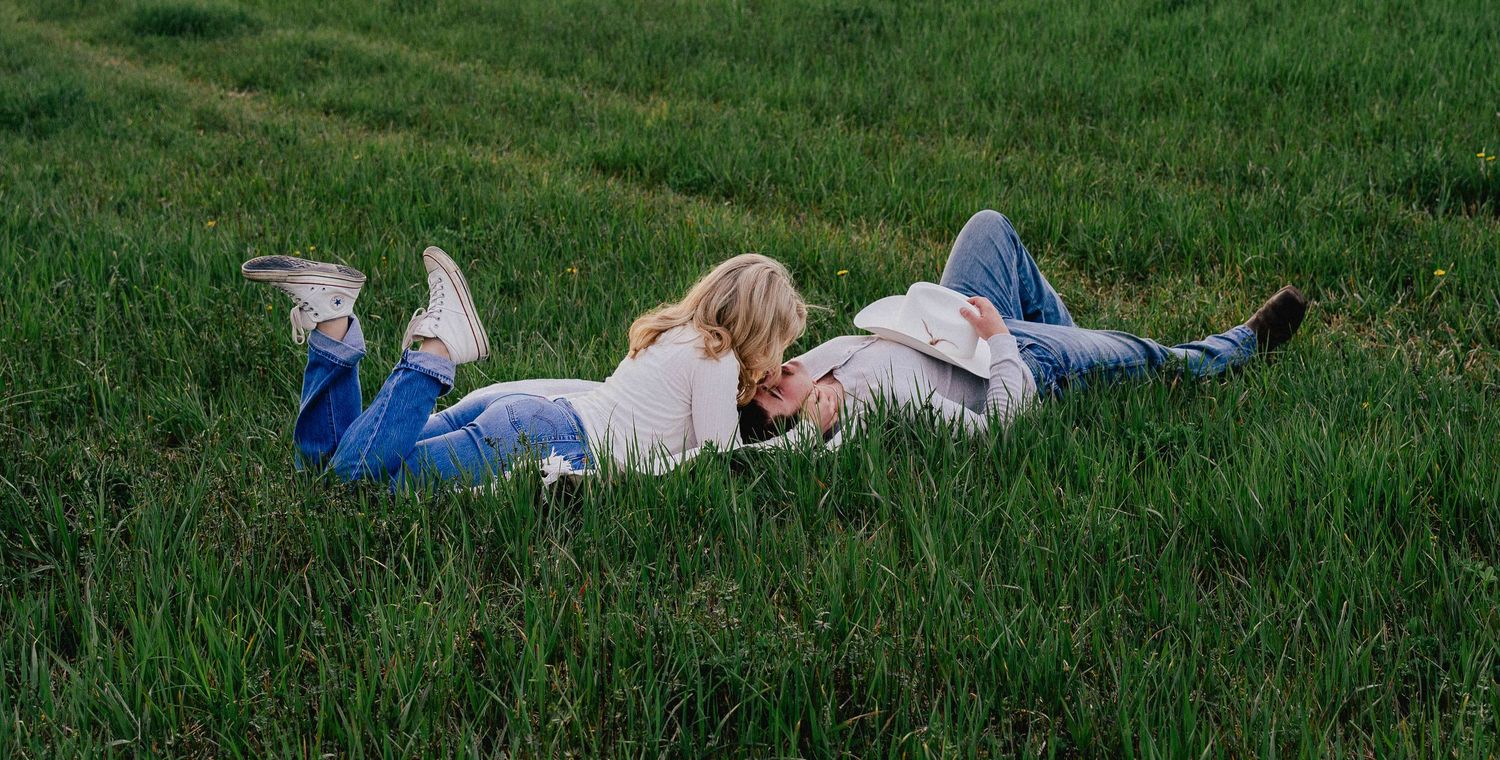 A couple lies together in a green grassy field, sharing a tender romantic moment outdoors.