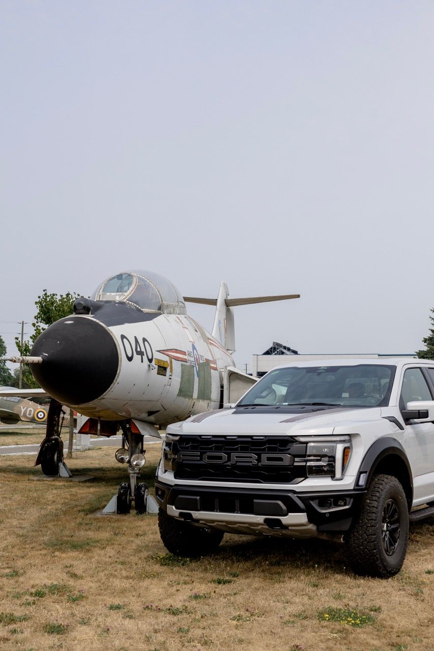 White Ford F-150 Raptor truck parked next to a vintage military jet fighter numbered 040 at an outdoor air museum.
