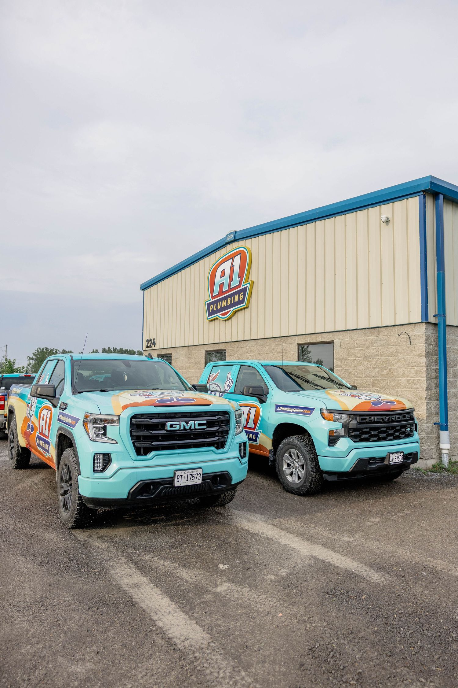 Two colorful Al Plumbing branded GMC and Chevrolet trucks parked outside the company building.