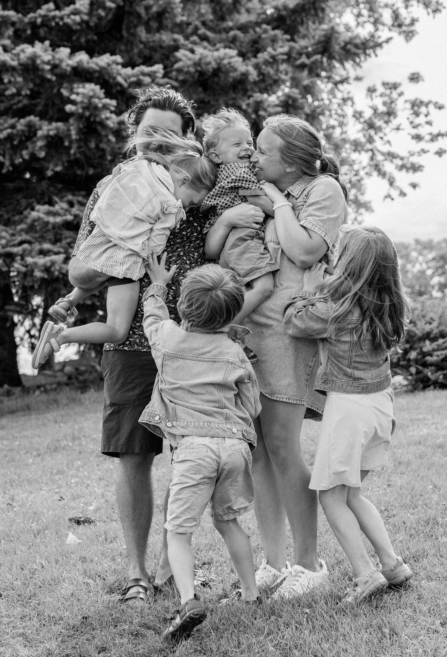 Black and white photo of a joyful family hugging and kissing outdoors on a grassy lawn with trees in the background.