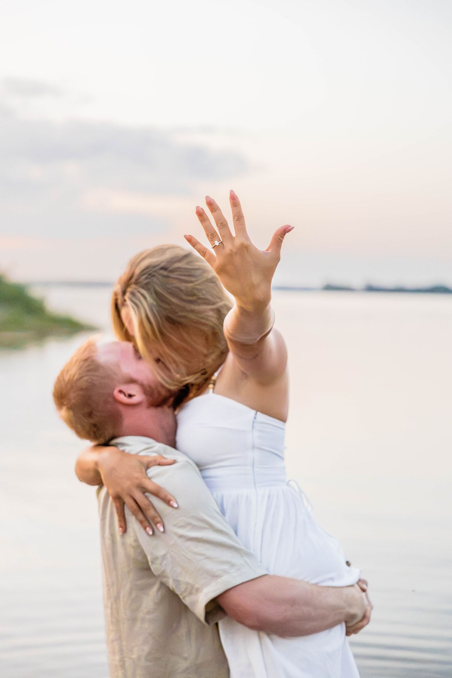 Couple embracing at sunset by the lake, woman showing off engagement ring while kissing her fiancé.