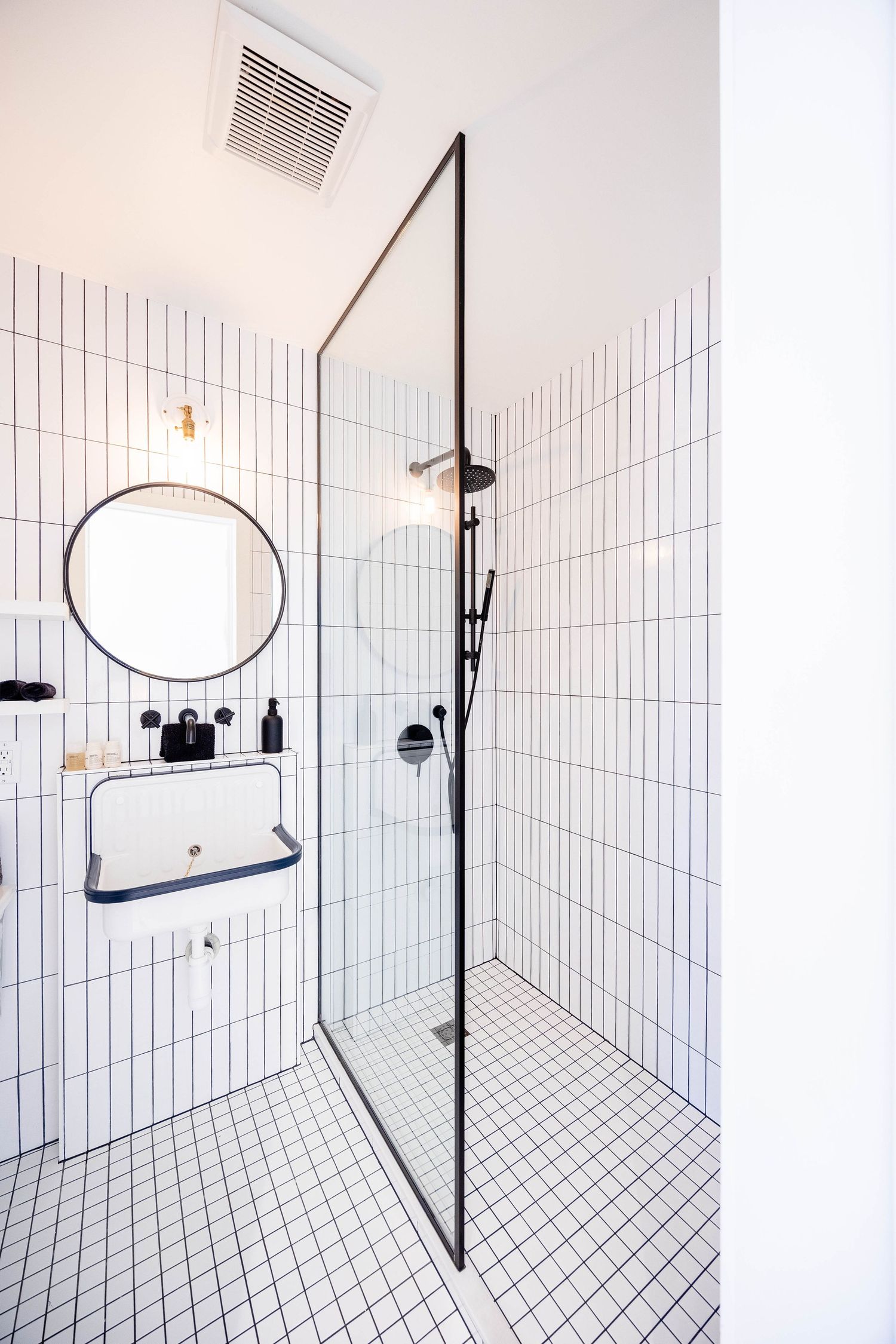 Modern white tiled bathroom with glass shower enclosure, round mirror, and black fixtures.