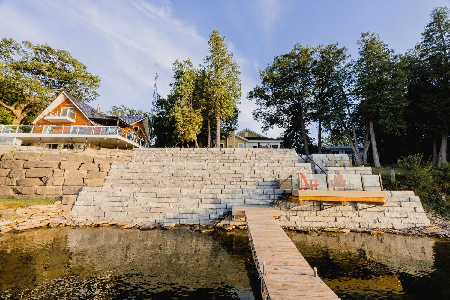 Stone retaining wall terraces leading to a wooden dock on a calm lake, with a red cottage and tall trees in background.