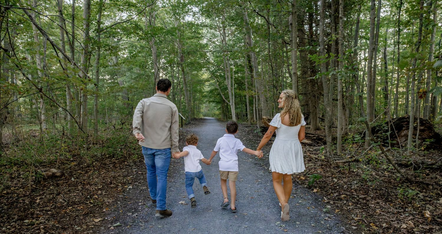 Family of four walking hand-in-hand down a lush forest path from behind.