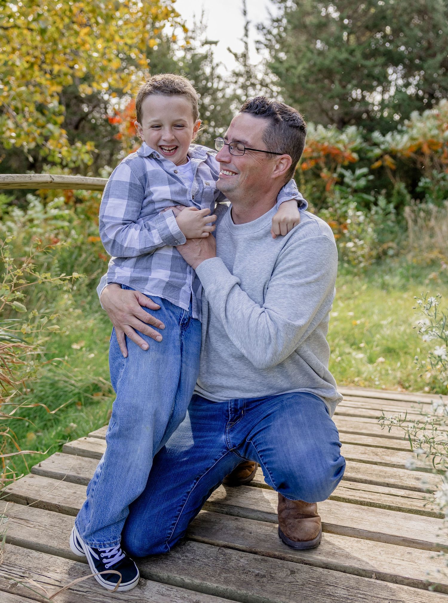 A father kneels on a wooden bridge, smiling at his laughing young son in a plaid shirt, surrounded by autumn foliage.