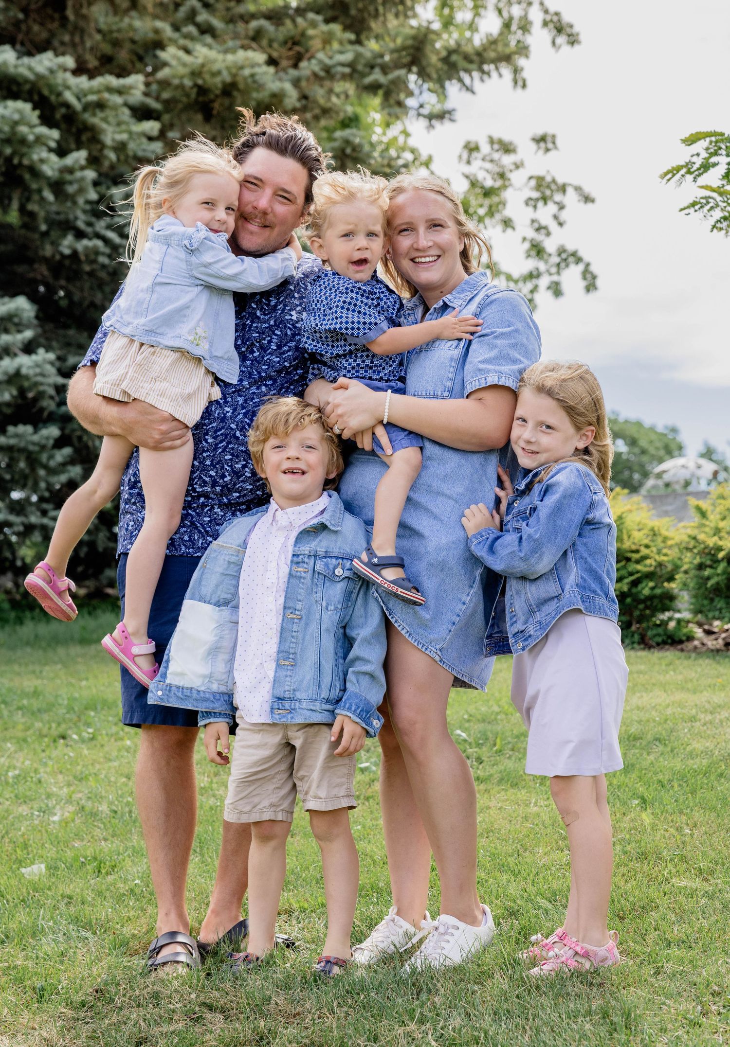 Happy family of six posing outdoors on green grass, parents holding young children, all wearing coordinating blue outfits.