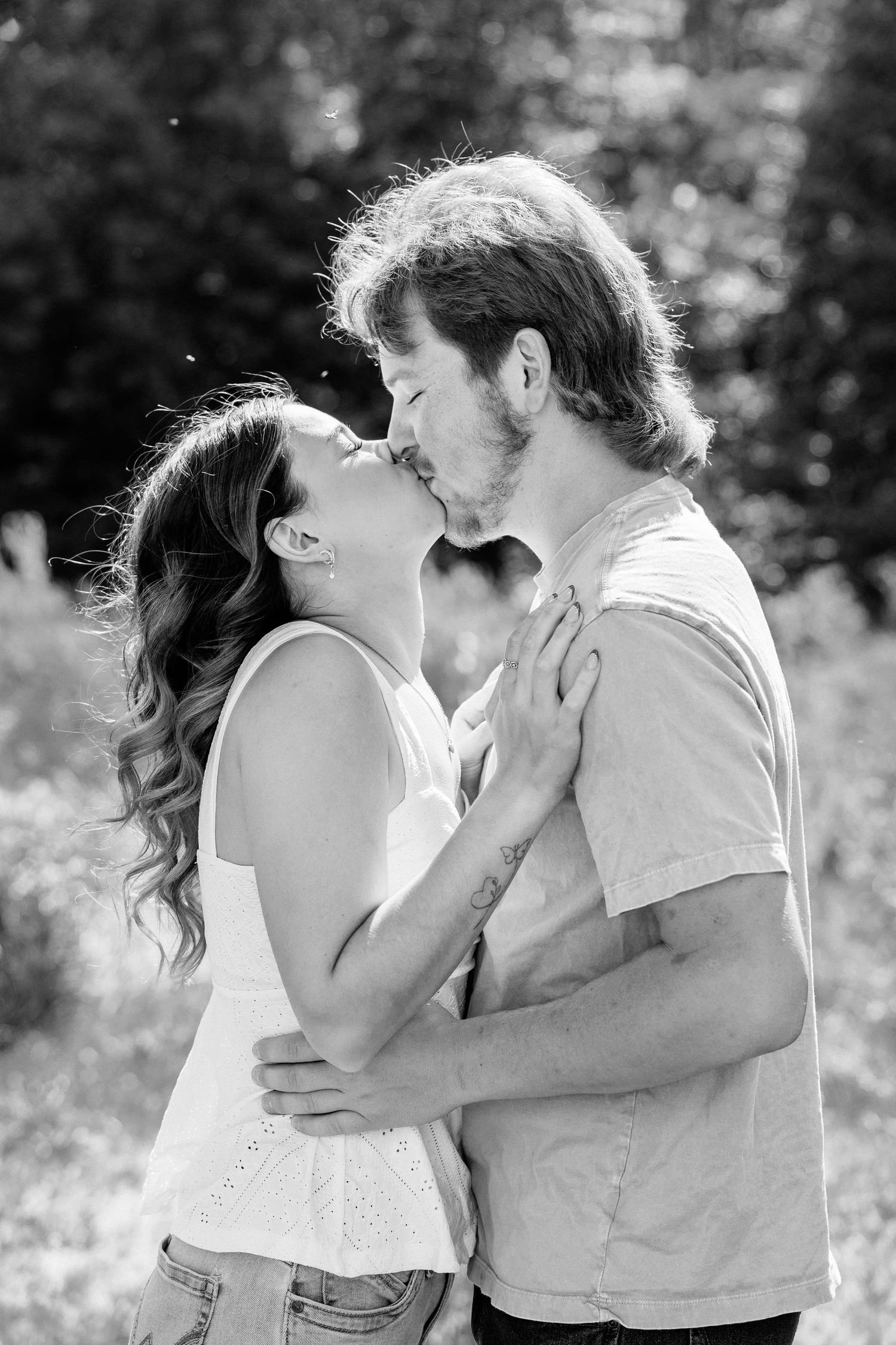 Black and white photo of a couple sharing a romantic kiss outdoors, embracing in a sunlit natural setting.