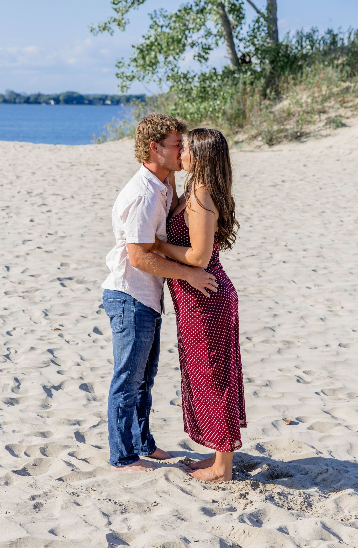 A couple shares a romantic kiss on a sandy beach, the woman in a red polka dot dress and the man in a white shirt and jeans.