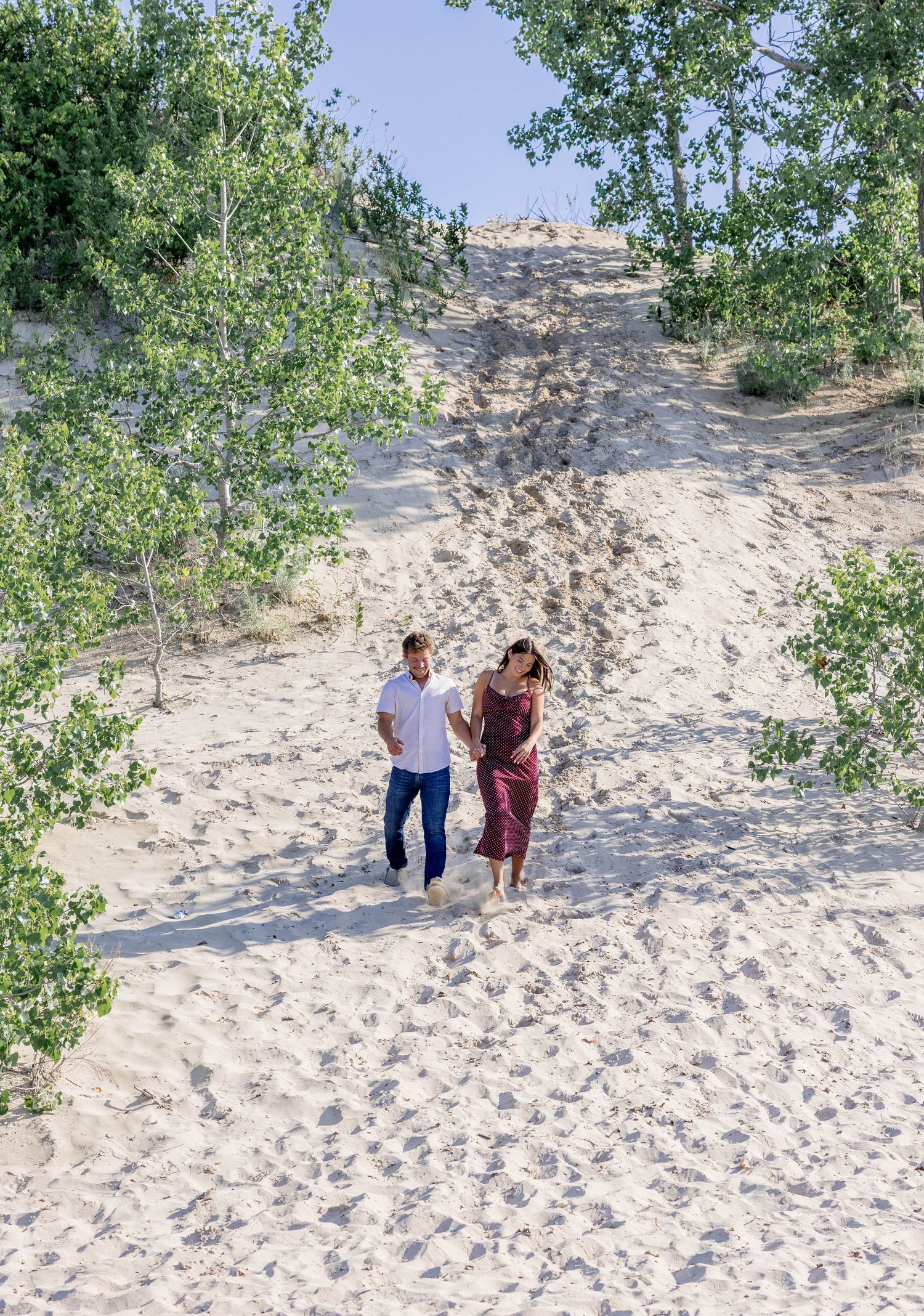 A couple walks hand-in-hand down sandy dunes surrounded by green trees on a sunny day.