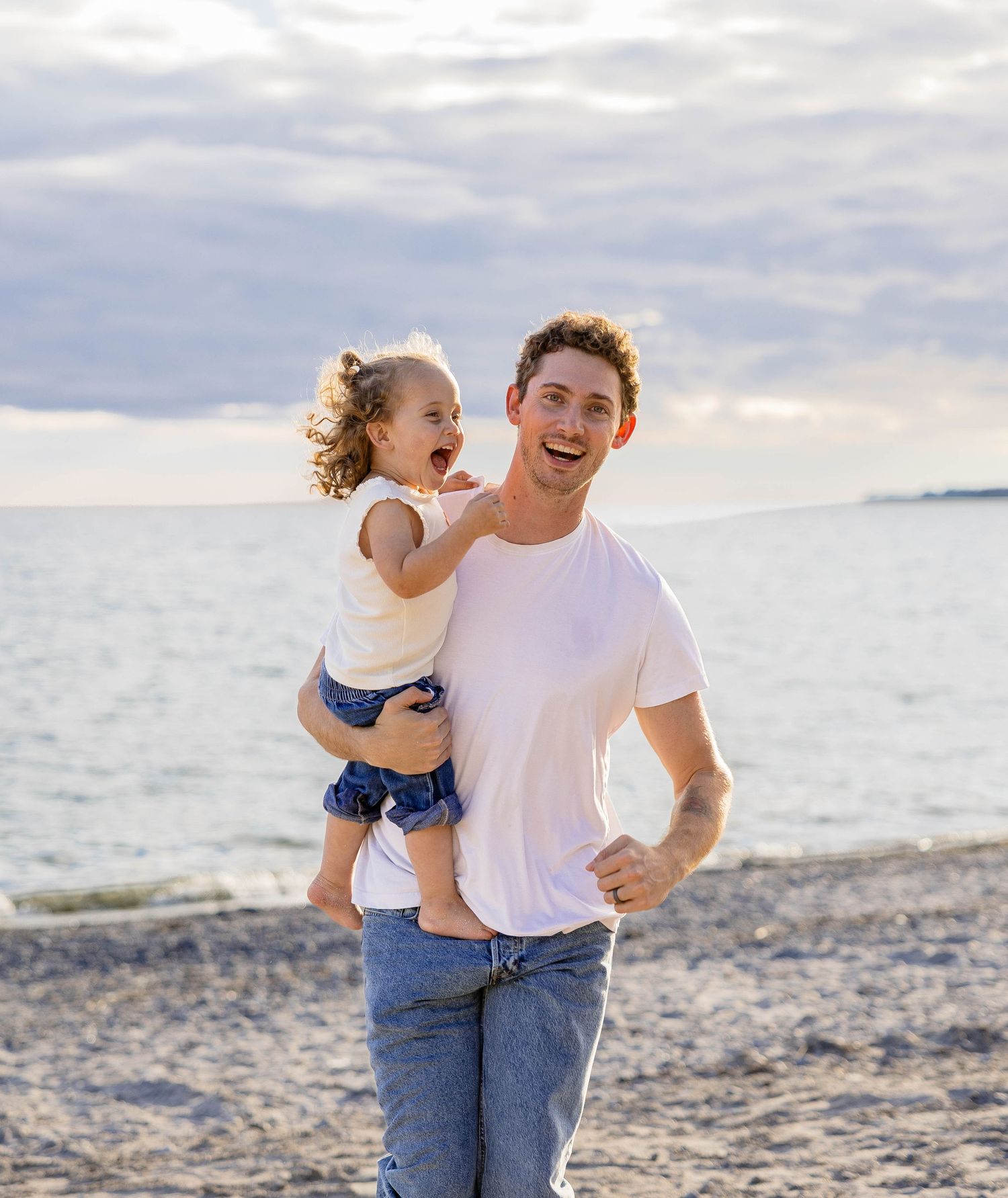 A smiling man in a white t-shirt holds a laughing toddler girl on a sandy beach under a cloudy sky.