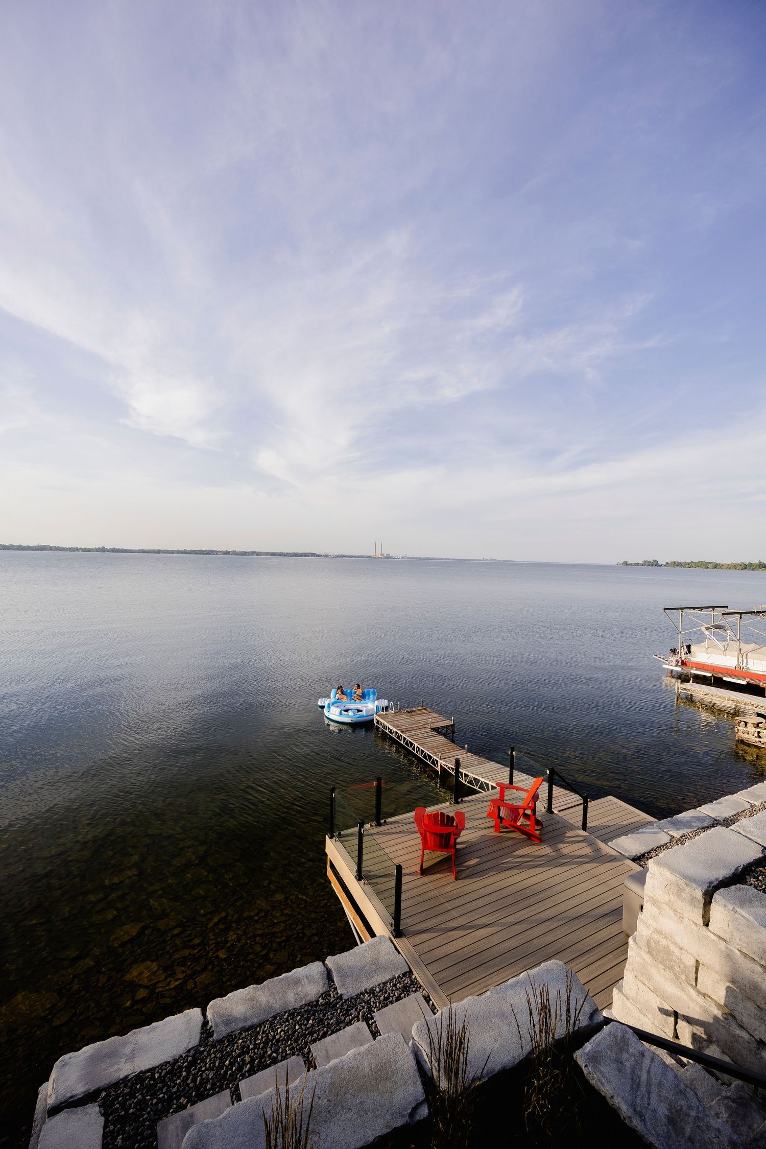 Wooden dock with red Adirondack chairs overlooking calm lake with paddleboat on sunny day.