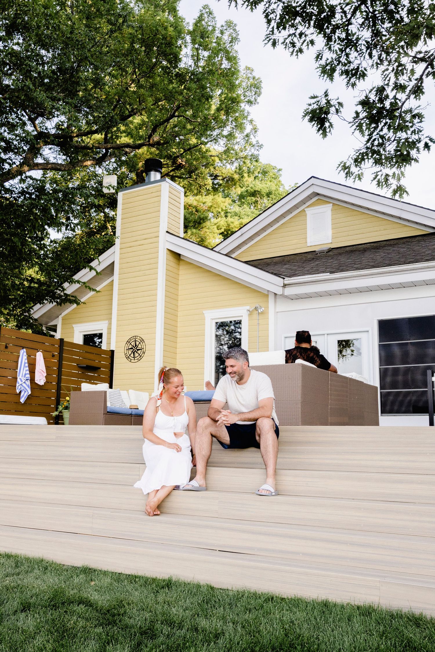 A couple sits on wooden deck steps outside a yellow house, enjoying a sunny day in a lush backyard.