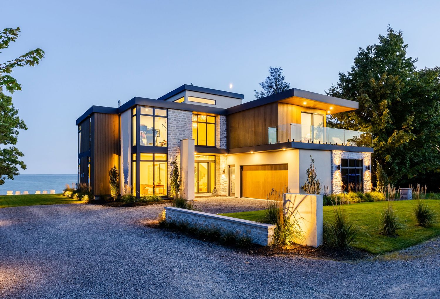 Contemporary two-story home with large windows, stone accents, and gravel driveway at twilight near the ocean.