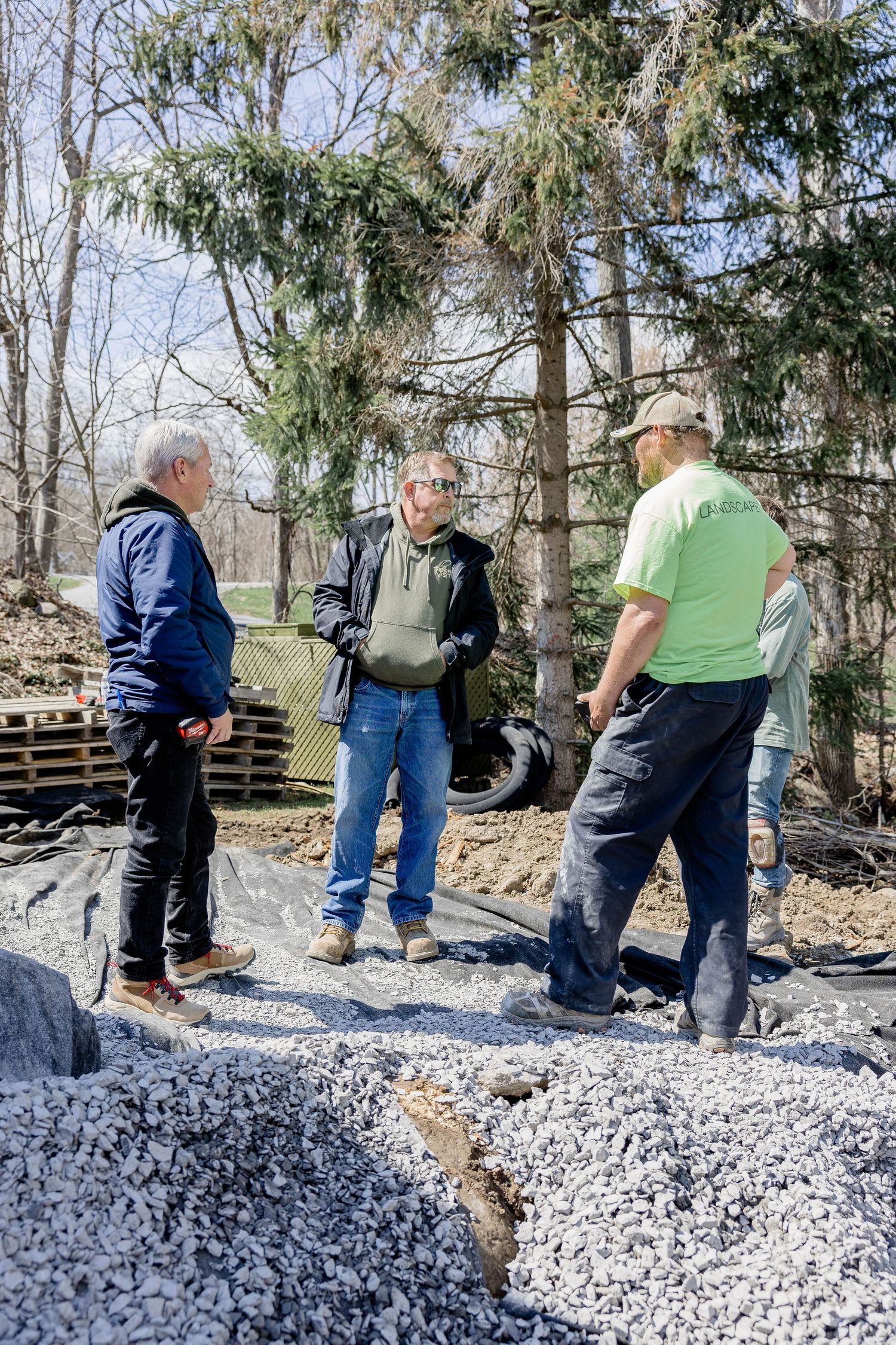 Three workers discuss project details while standing on a gravel surface at an outdoor construction site.