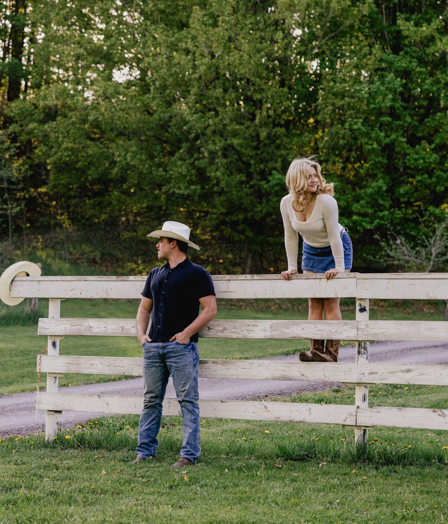 Cowboy in white hat stands by white fence as blonde woman sits on top smiling.