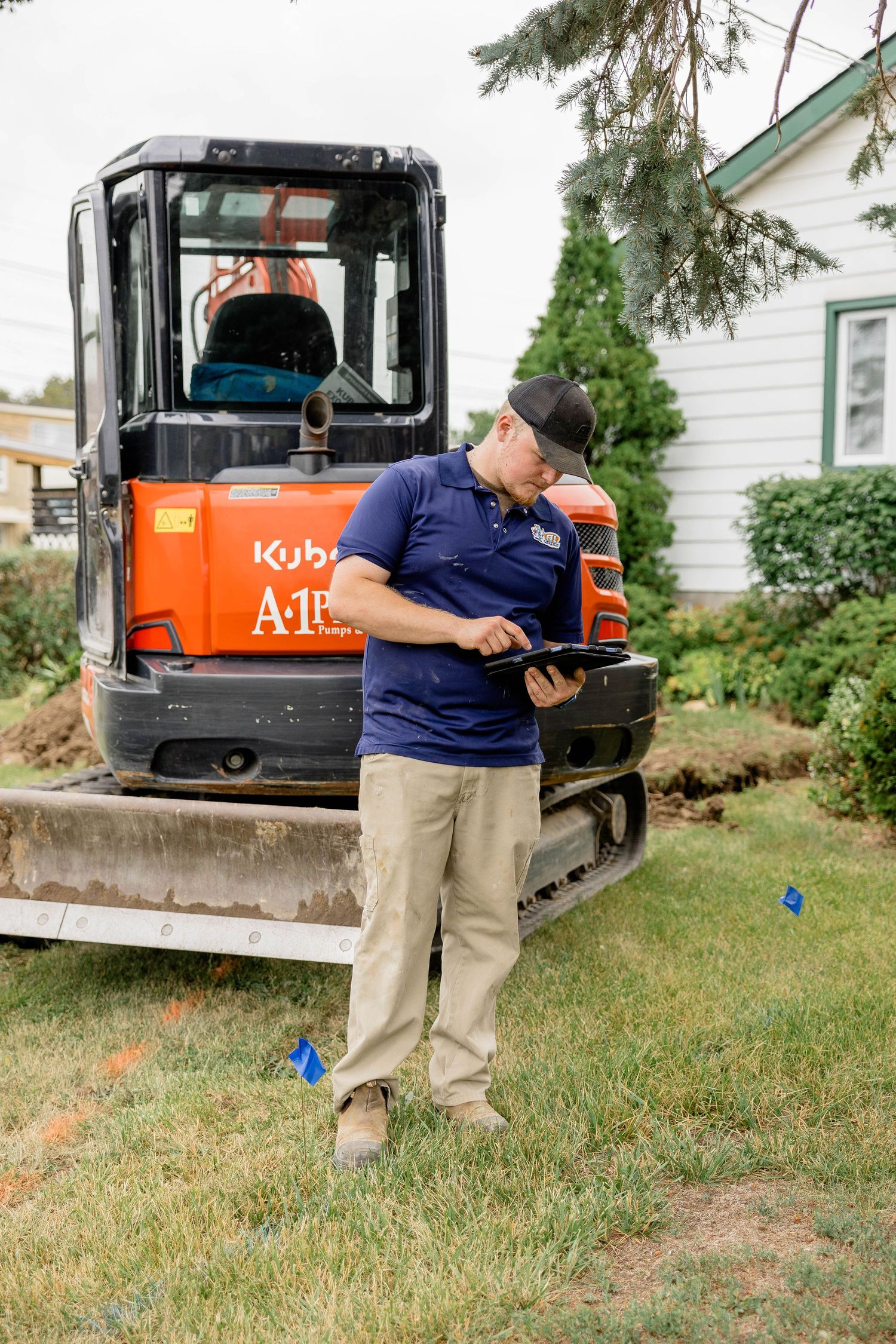 A technician in a blue polo reviews a tablet near a Kubota mini excavator on a residential lawn.
