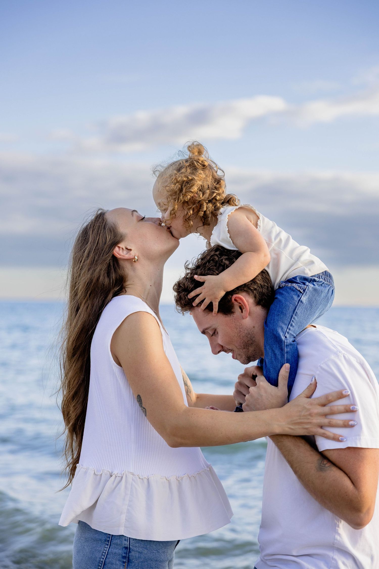 Mom kissing toddler on dad's shoulders at beach, family enjoying sunny day by the ocean.