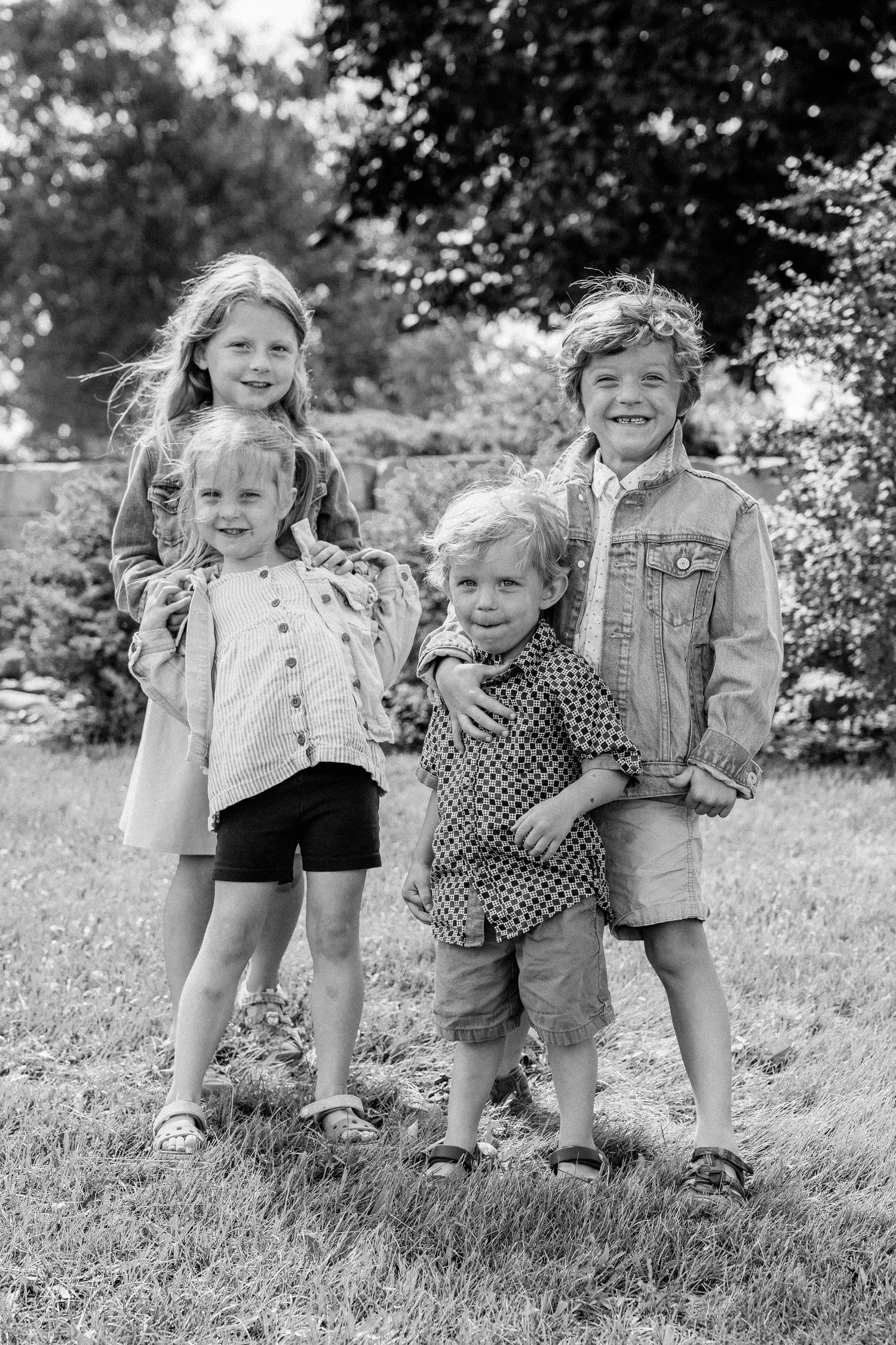 Black and white photo of four young children smiling together outdoors on a grassy lawn with trees in the background.