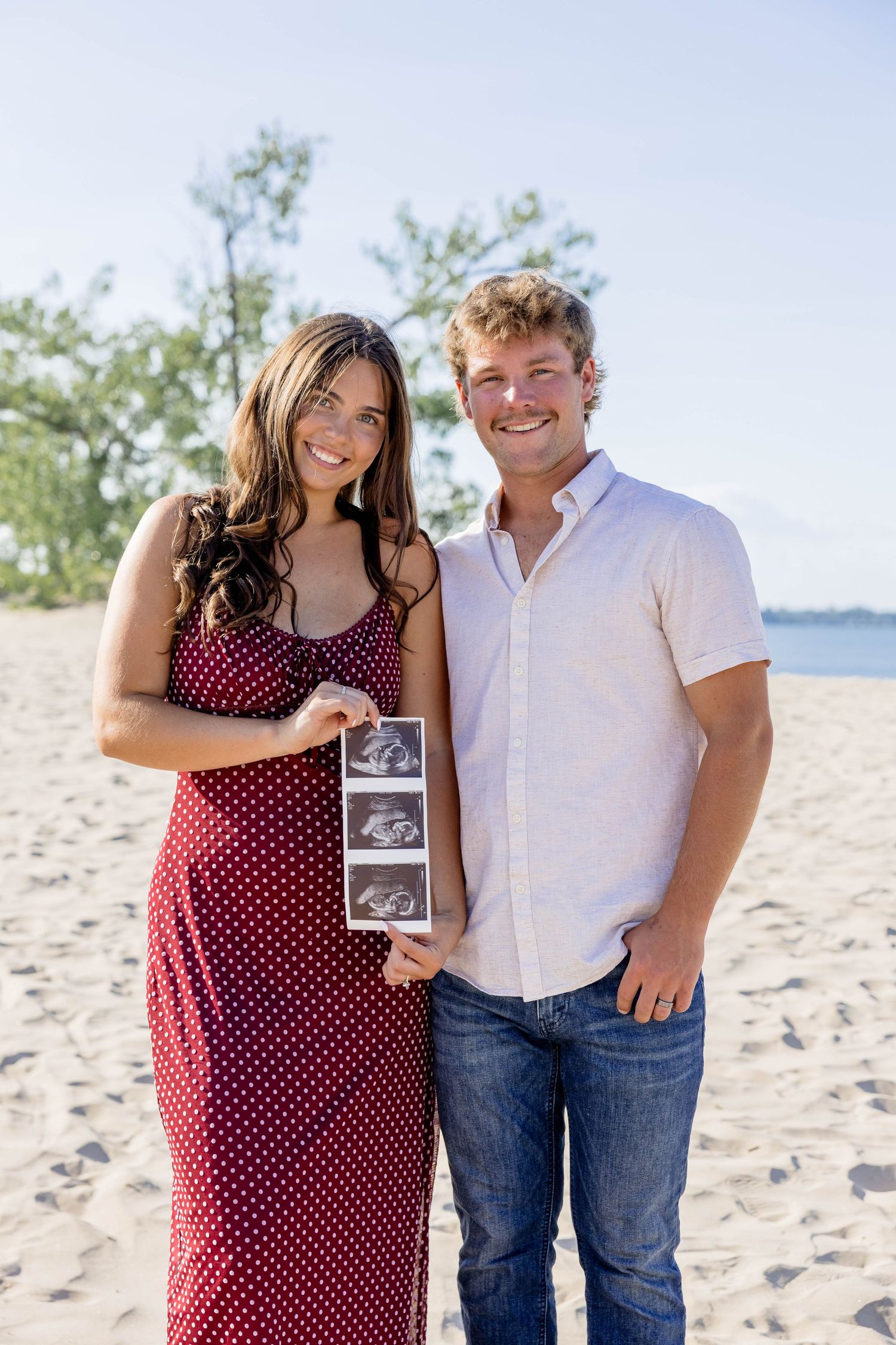 Happy couple smiling on sandy beach holding ultrasound photos announcing pregnancy, woman in red polka dot dress.